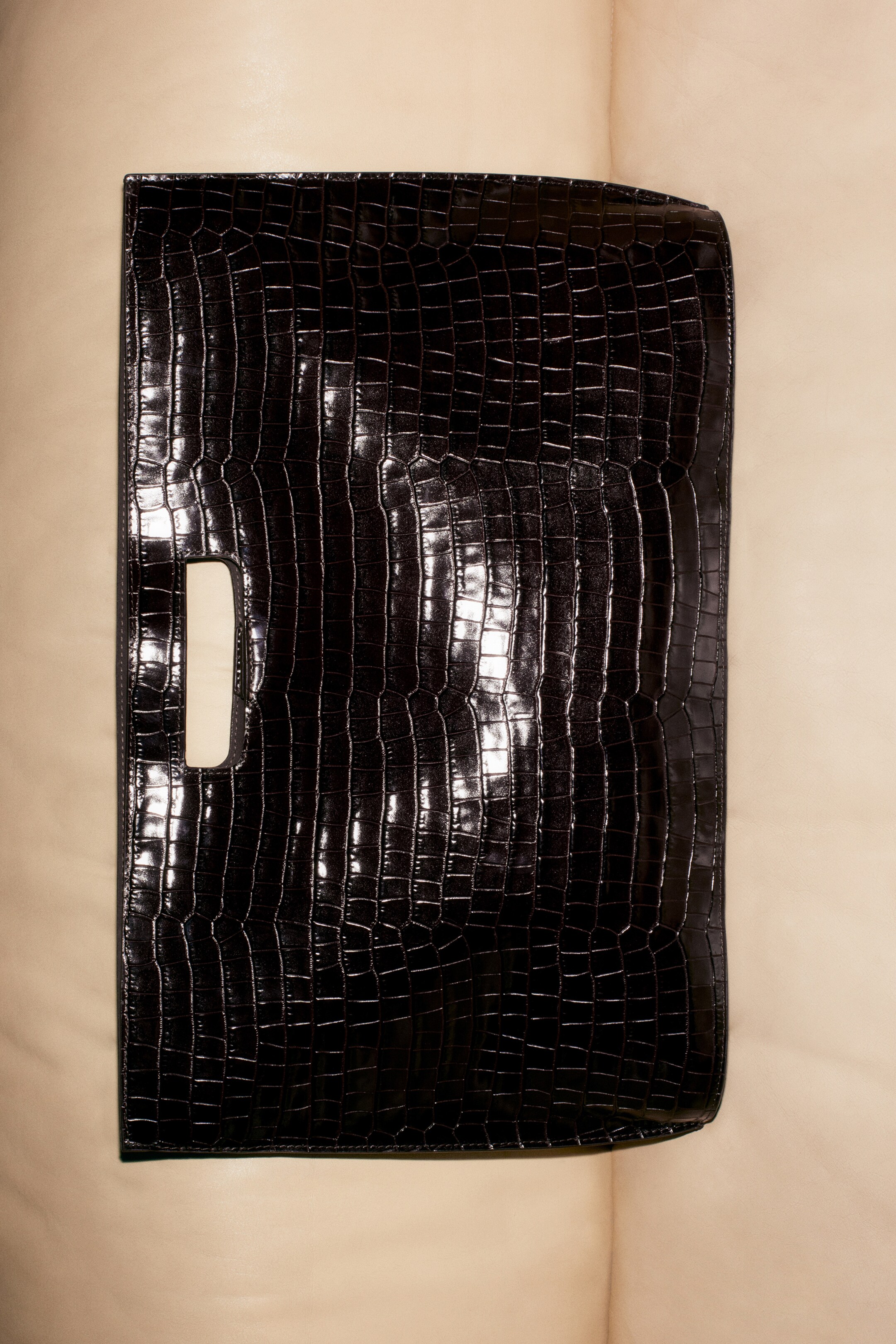 Größeres Bild anzeigen: Eine schwarze Folio-Tasche liegt flach und wird aus einer Vorderansicht präsentiert. Sie zeichnet sich durch eine glänzende, krokodilartige Textur über ihre rechteckige Form mit dezent abgerundeten oberen und unteren Ecken aus und hat einen rechteckigen Ausschnittgriff auf ihrer linken Seite.