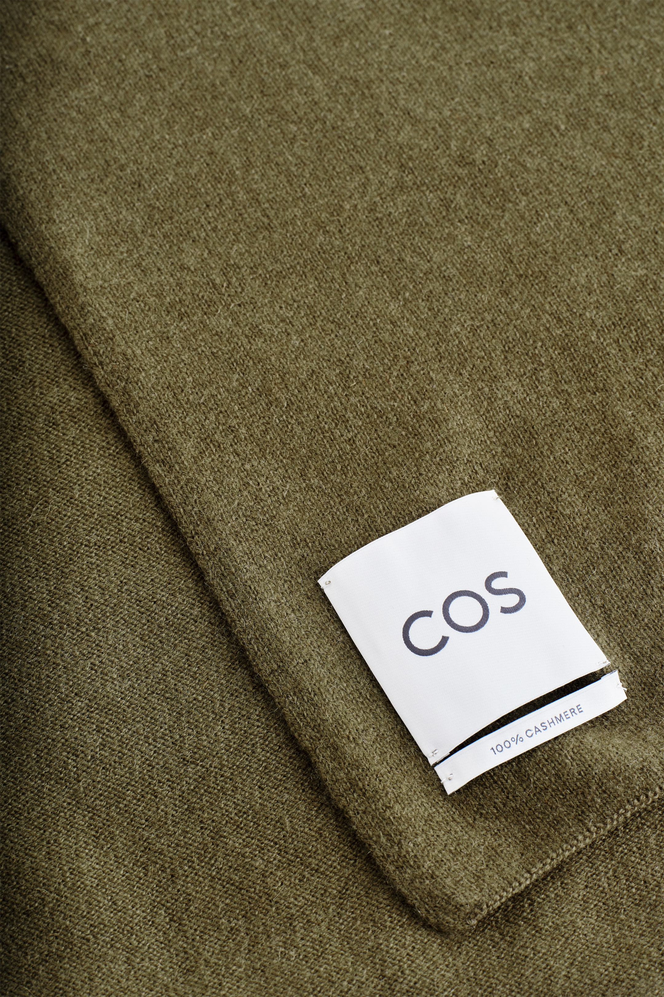 Größeres Bild anzeigen: Ein olivgrüner Schal zeigt eine fein gestrickte Textur. Ein weißes Etikett, auf dem 'COS' und '100% CASHMERE' steht, ist am Stoff angebracht.