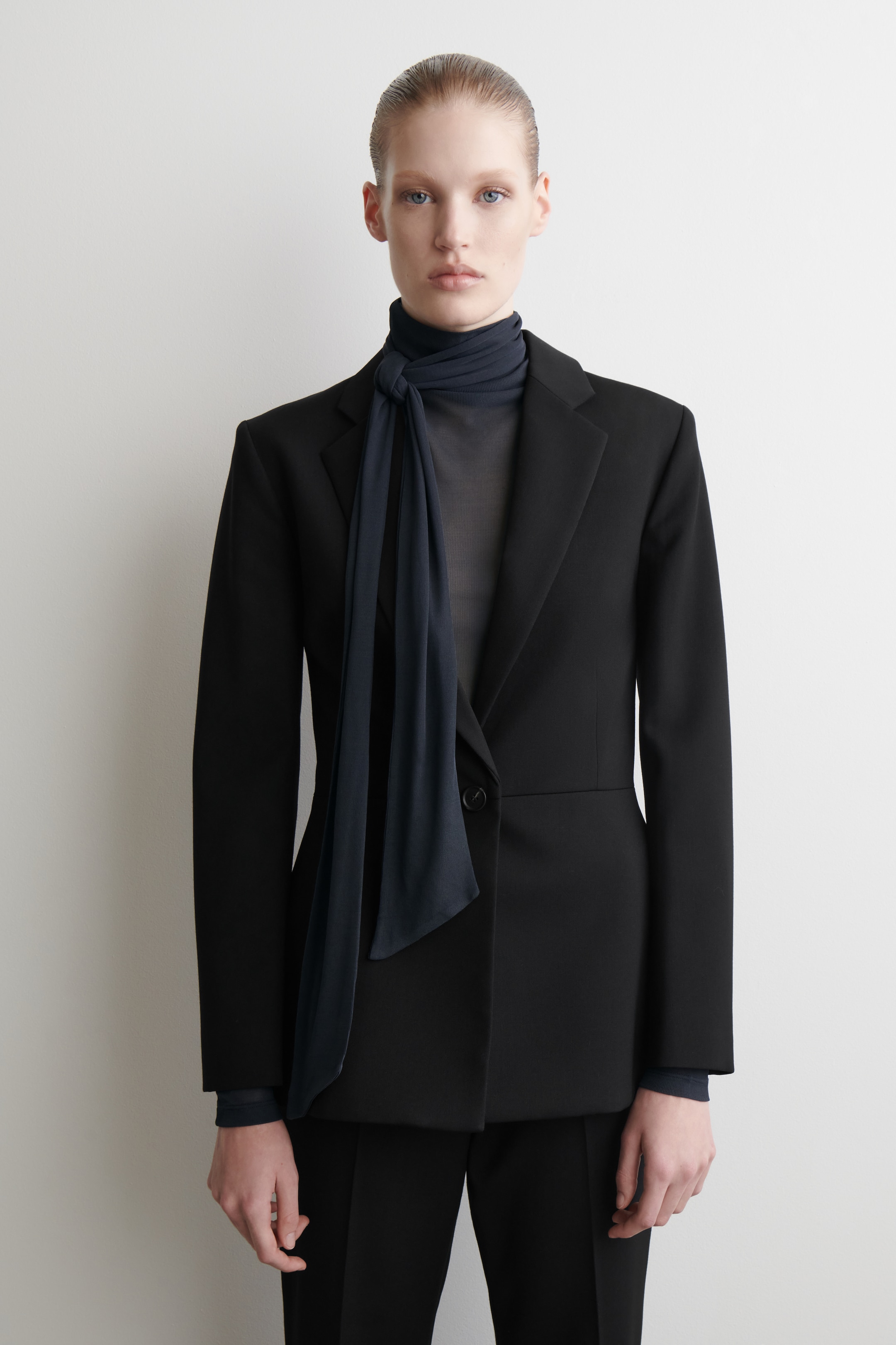 Größeres Bild anzeigen: Ein dunkelblaues, halbtransparentes Oberteil wird VORNE gezeigt, unter einem schwarzen Blazer getragen, mit einem hohen Kragen und einem passenden Schal, der um den Hals gebunden und vorne herab drapiert ist.