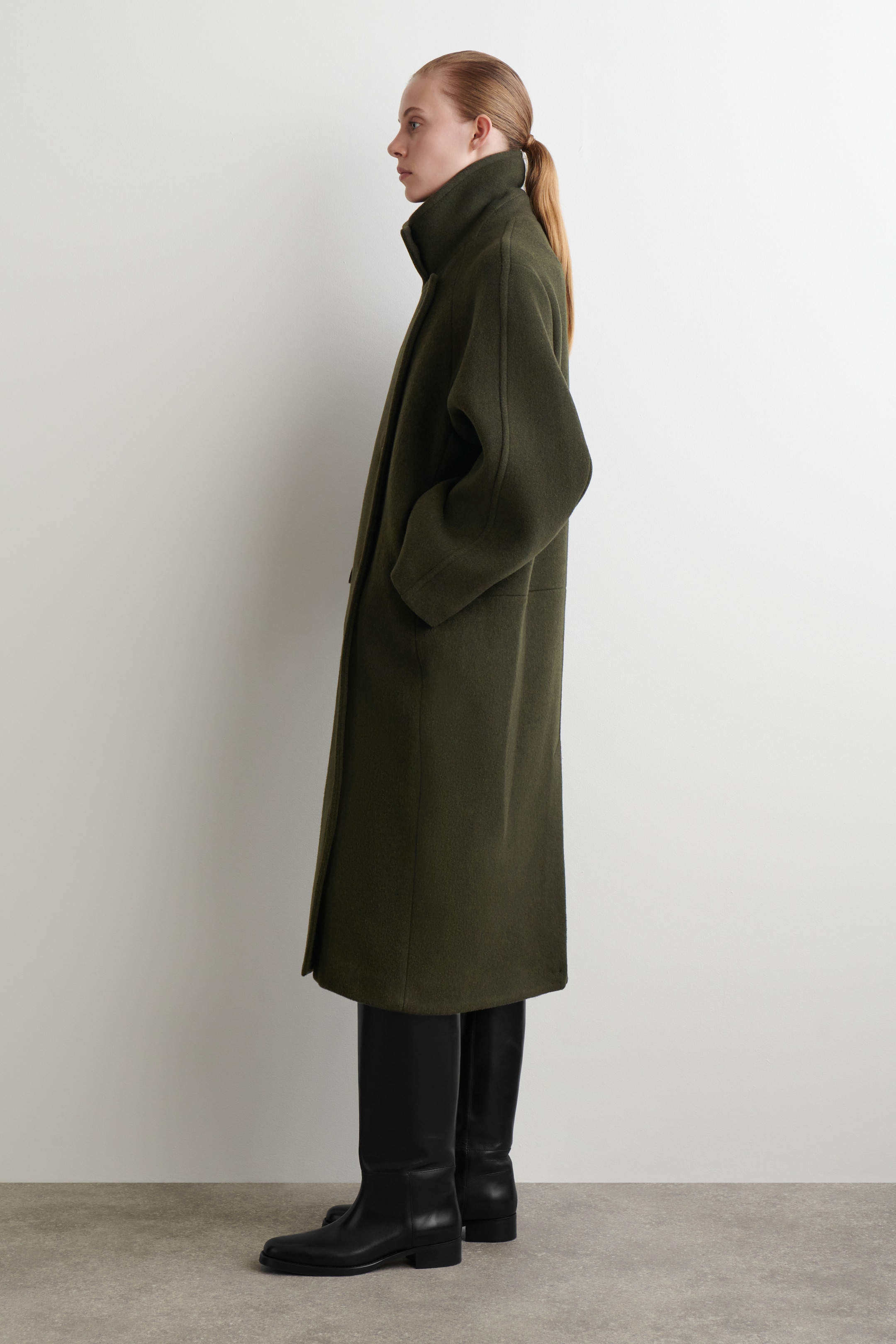 Visualizza immagine più grande: Un cappotto verde oliva di lunghezza media al polpaccio viene presentato di profilo, mettendo in mostra la sua vestibilità rilassata con spalle cadenti e un collo a imbuto alto. Il cappotto presenta tasche laterali sottili e una texture liscia e infeltrita.