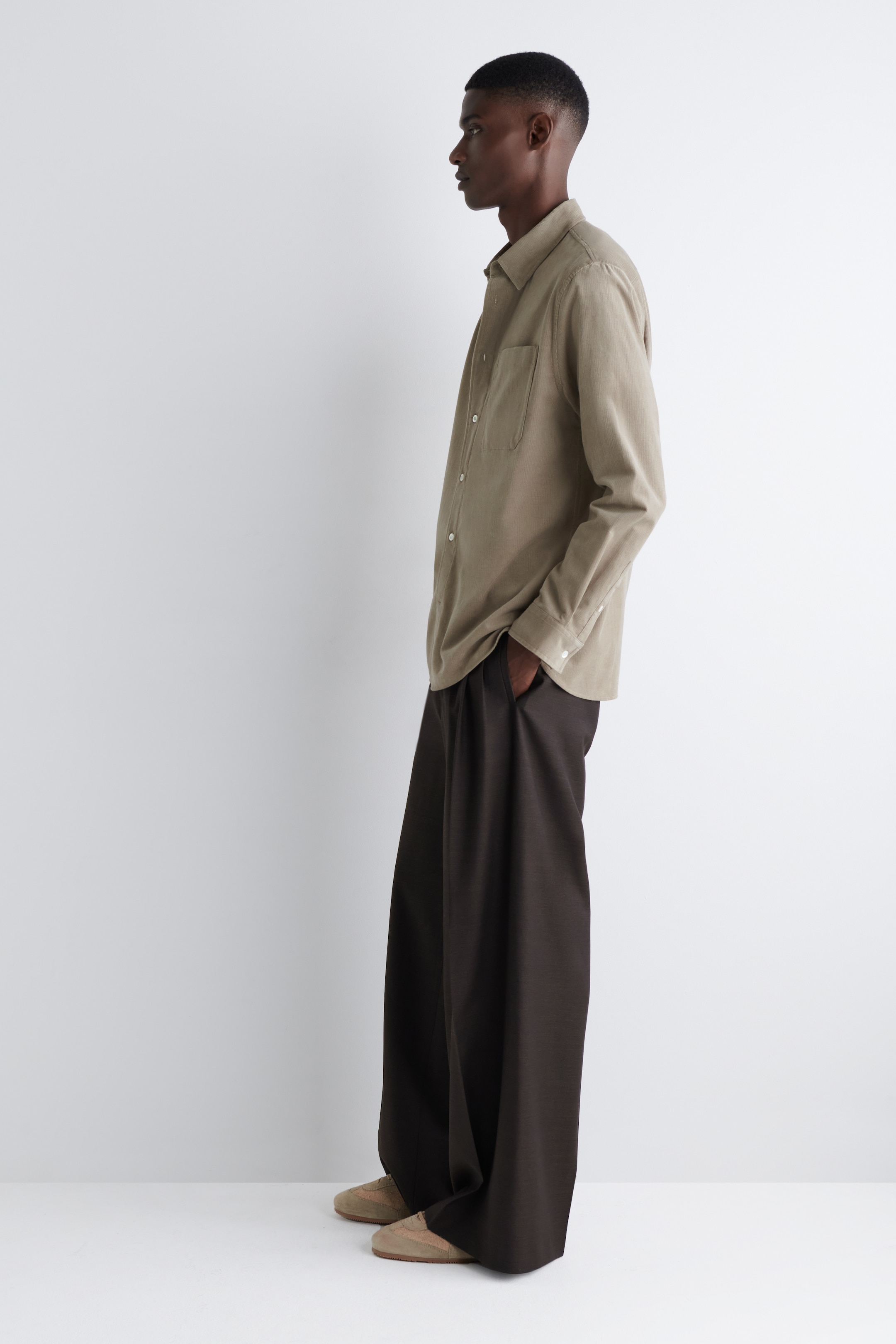 Agrandir l'image: Une chemise boutonnée à manches longues de couleur brun clair, avec une poche poitrine, est portée non rentrée sur un pantalon large de couleur brun foncé, présentée de profil. La coupe décontractée de la chemise et le drapé ample du pantalon, qui s'accumule sur des chaussures basses de couleur beige clair, sont visibles.