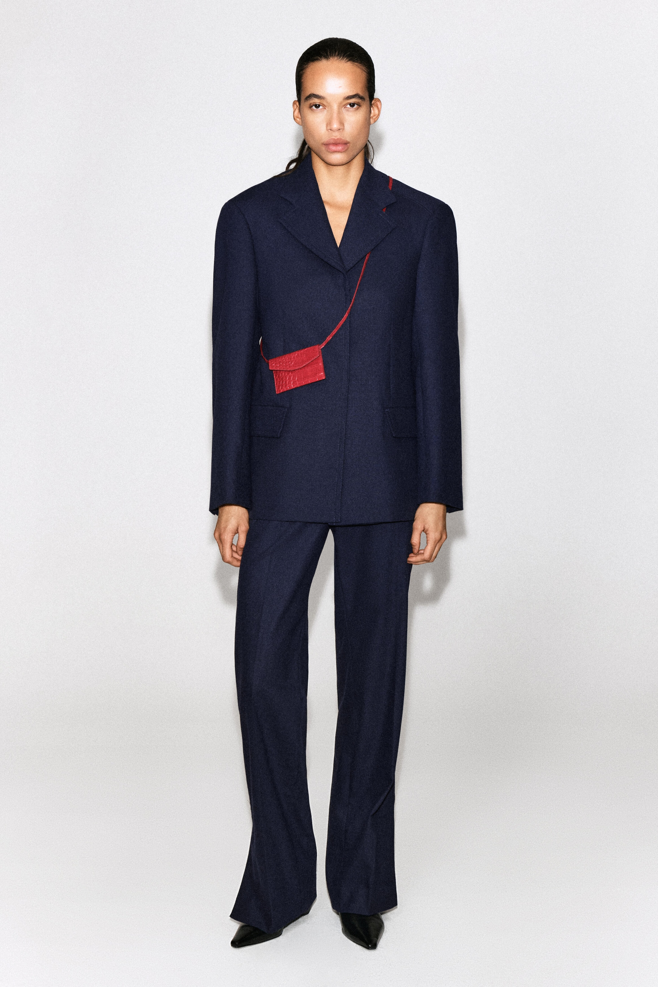 A red leather card holder with a crocodile-embossed texture is shown from the front, worn crossbody over a navy blue blazer. The small rectangular card holder has an envelope-style flap and hangs from a thin, matching red strap slung diagonally across the torso. A red leather card holder with a crocodile-embossed texture is shown from the front, worn crossbody over a navy blue blazer. The small rectangular card holder has an envelope-style flap and hangs from a thin, matching red strap slung diagonally across the torso.
