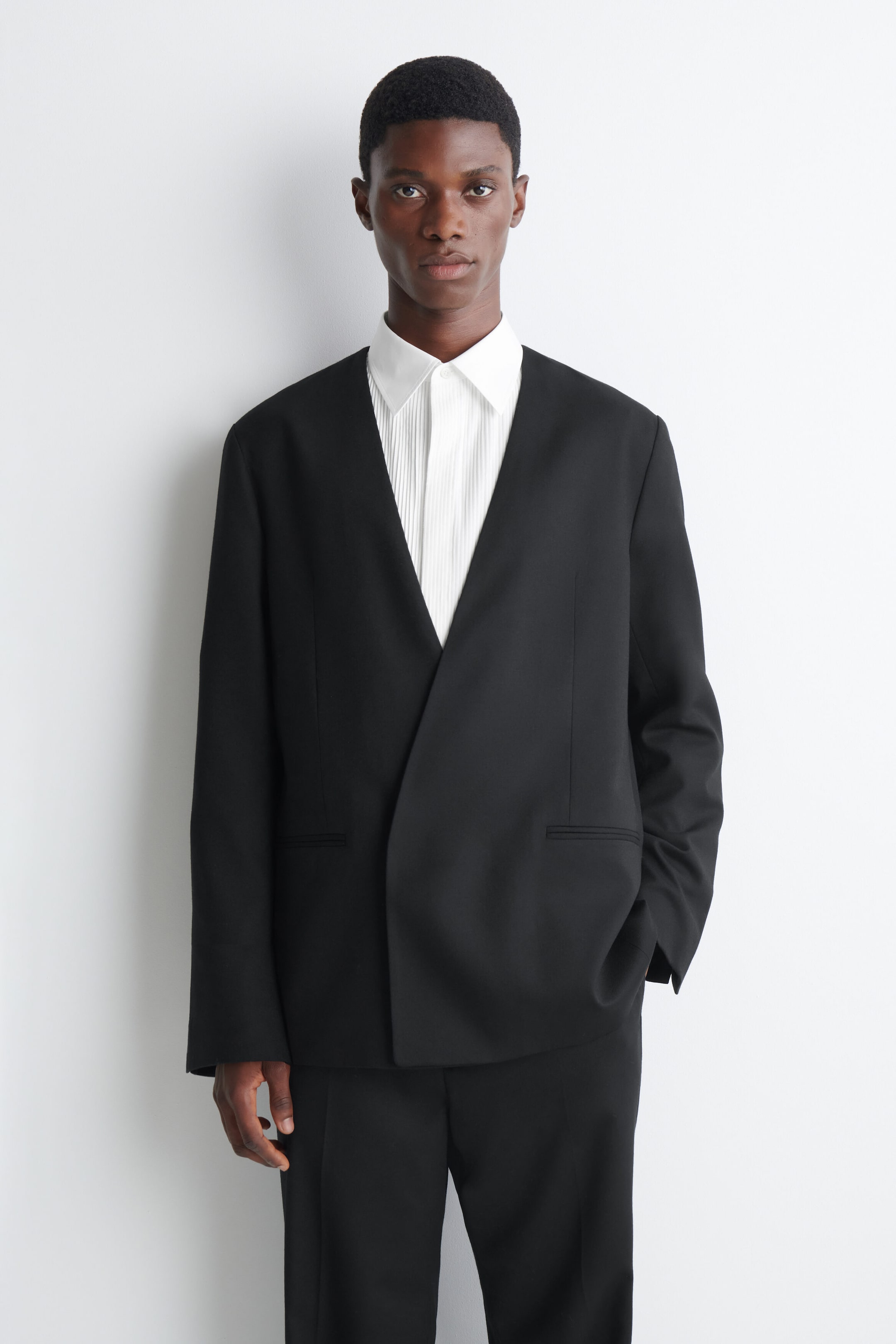 A collarless black blazer featuring a deep V-neckline and a clean wrap-front design is worn by the model. The blazer is layered over a white pleated-front collared shirt and paired with matching black trousers. A collarless black blazer featuring a deep V-neckline and a clean wrap-front design is worn by the model. The blazer is layered over a white pleated-front collared shirt and paired with matching black trousers.