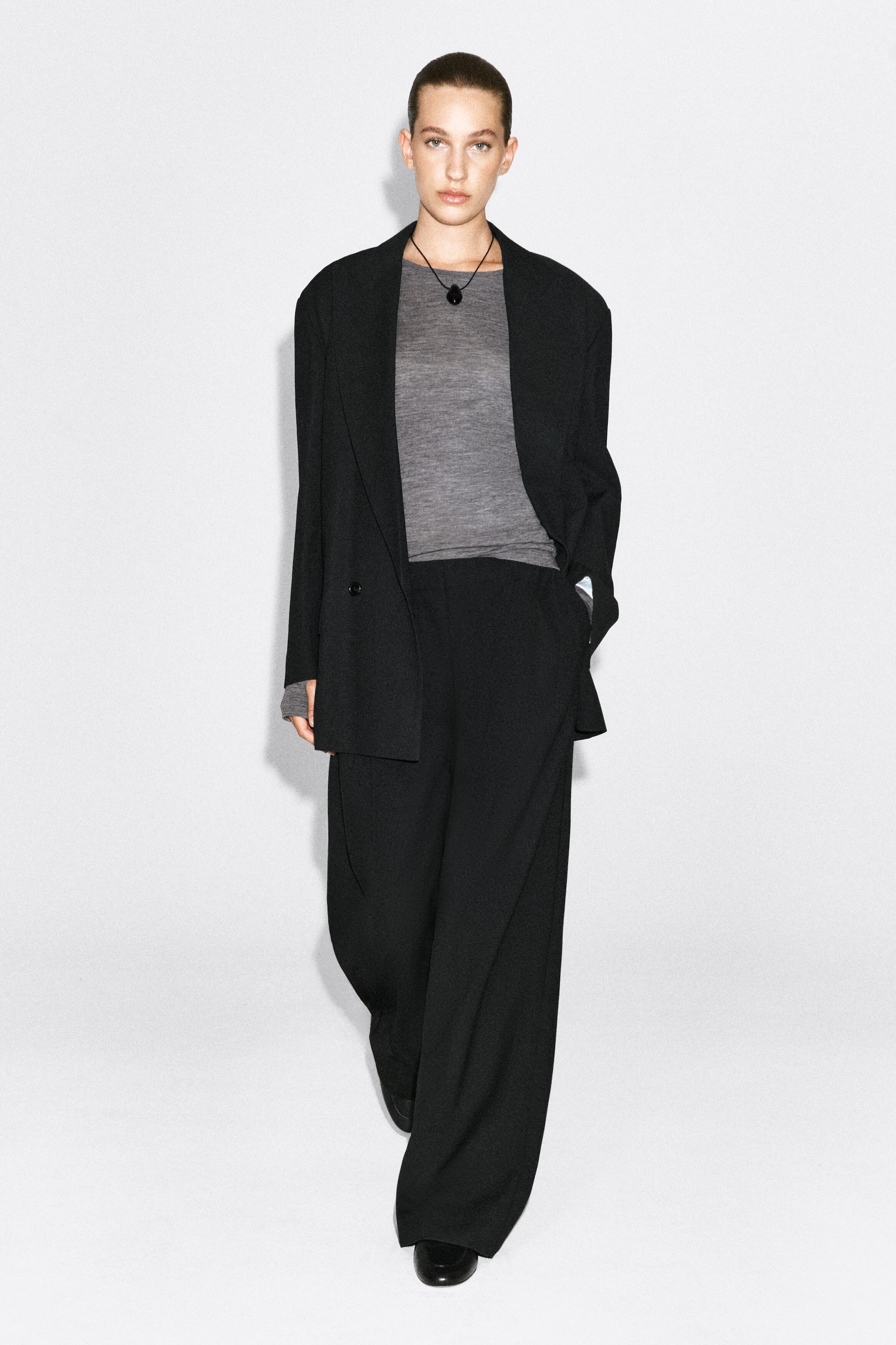 A grey, slim-fit merino wool top with a crew neck is being worn by the model as a base layer. The long-sleeve top is styled underneath an open black blazer and tucked into black wide-leg trousers.