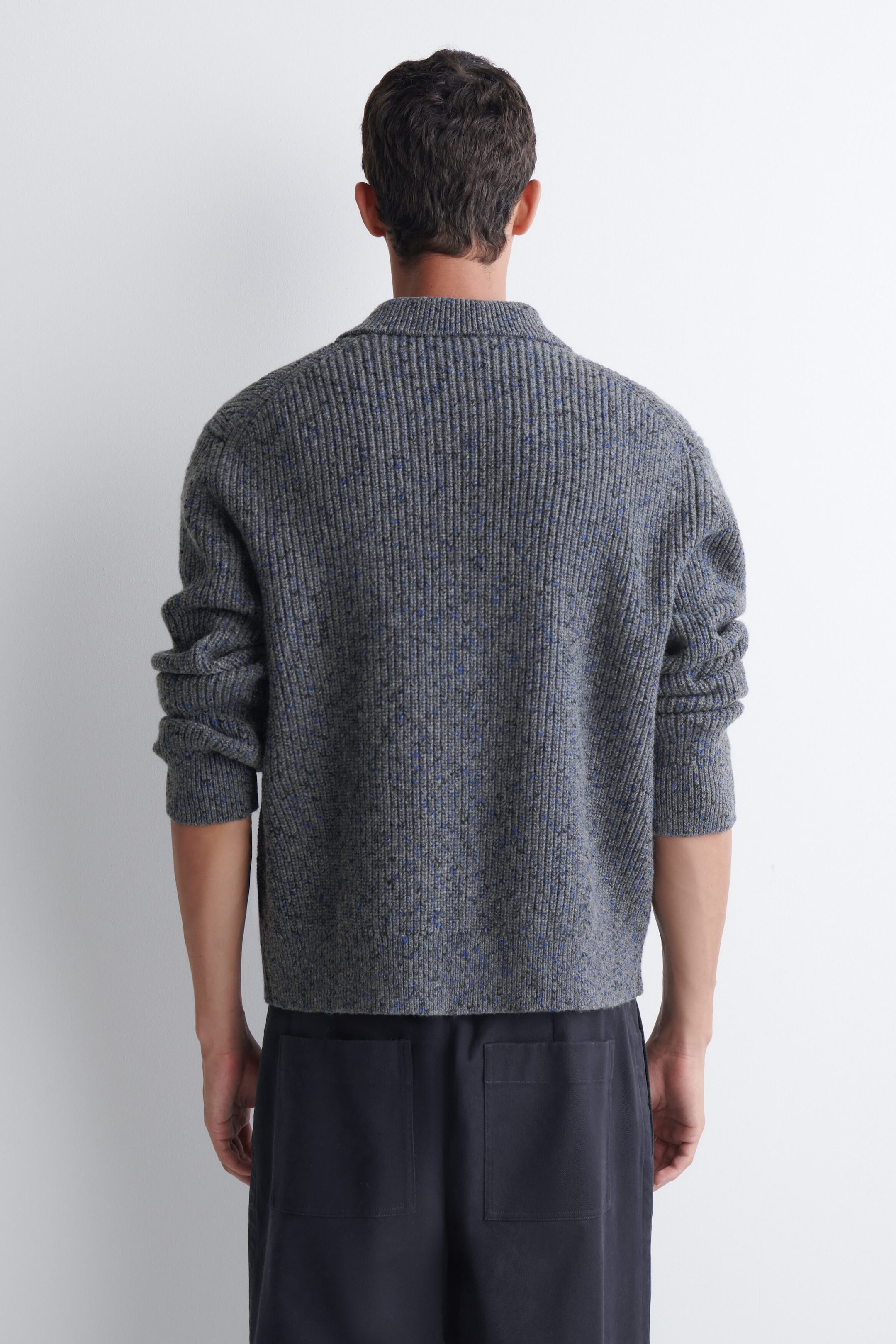 A grey ribbed-knit sweater with blue speckled details is being worn by the model, shown from the back. The sweater features a high ribbed collar and long sleeves pushed up slightly, and is paired with dark trousers. A grey ribbed-knit sweater with blue speckled details is being worn by the model, shown from the back. The sweater features a high ribbed collar and long sleeves pushed up slightly, and is paired with dark trousers.