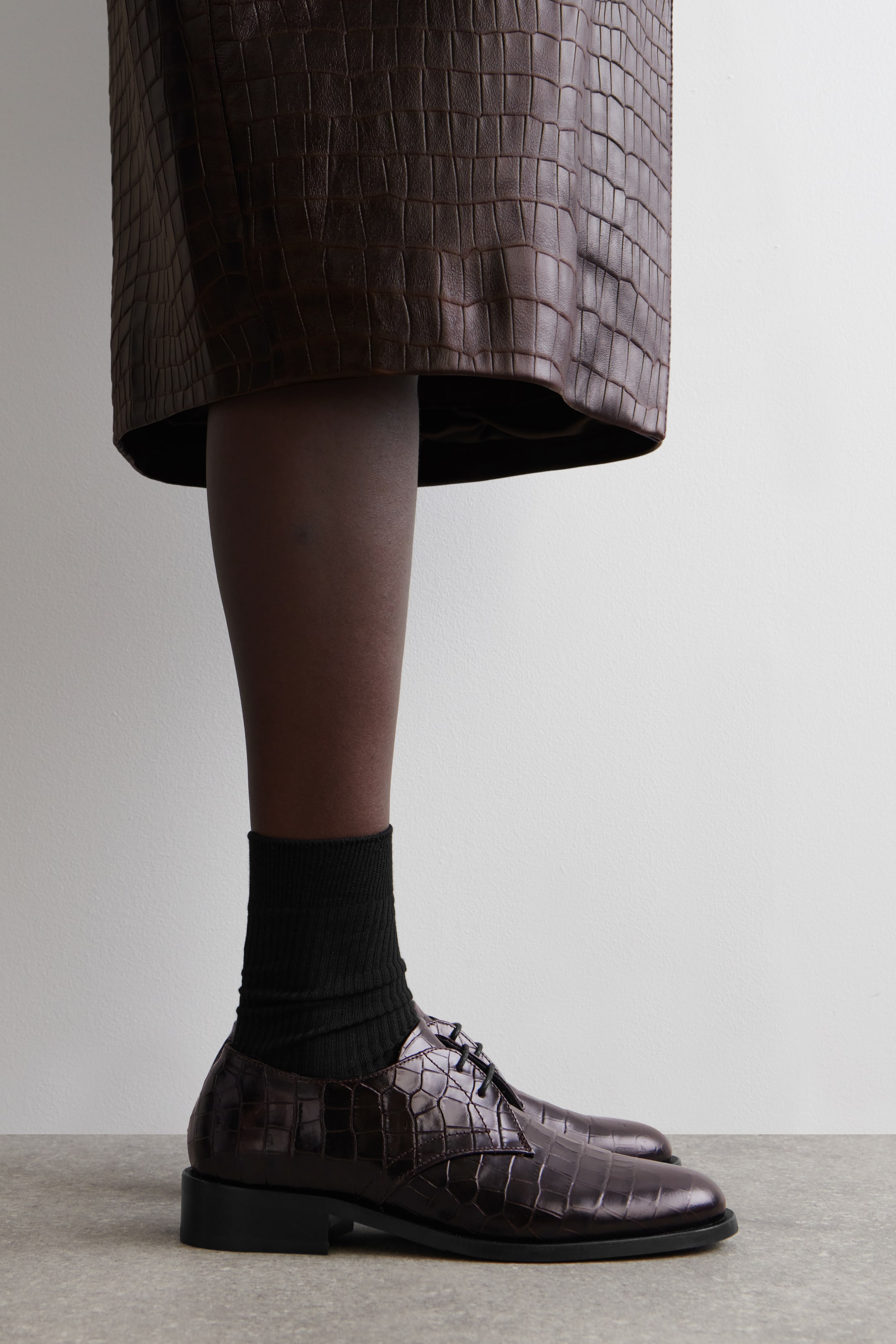 Grösseres Bild ansehen: Ein Espresso-farbener Derby-Schuh aus Krokodil-geprägtem Leder zeigt nach rechts, mit einer abgerundeten Spitze, schwarzen gewachsten Baumwollschnürsenkeln und einer niedrigen, flachen schwarzen Sohle.