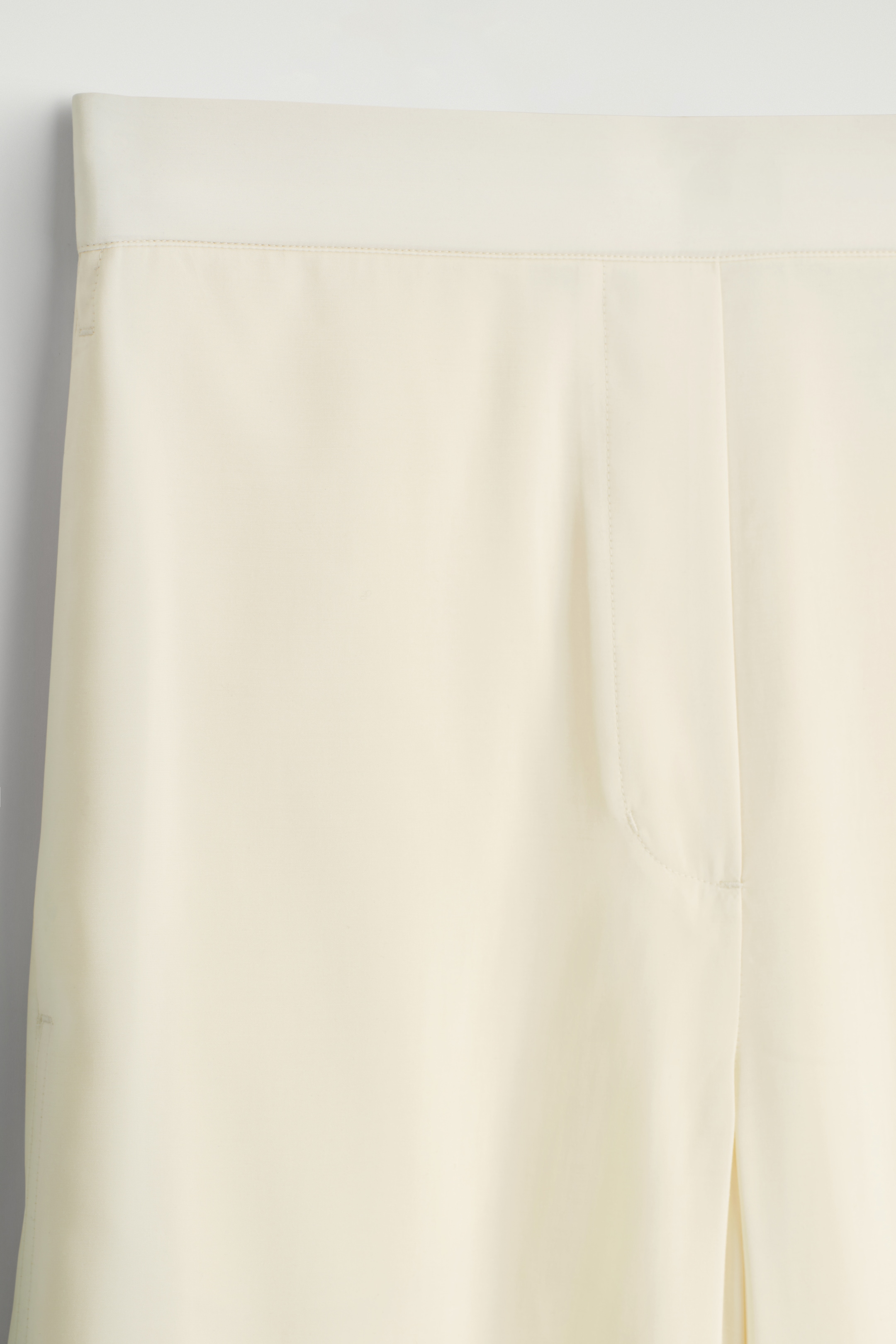 Agrandir l'image: Un pantalon crème est exposé de face, montrant leur partie supérieure. Ils présentent une large ceinture plate avec des surpiqûres subtiles, et leur tissu lisse reflète un éclat doux. Une ligne courbe légère sur la gauche suggère une ouverture de poche, et une couture verticale descend le long du côté droit.