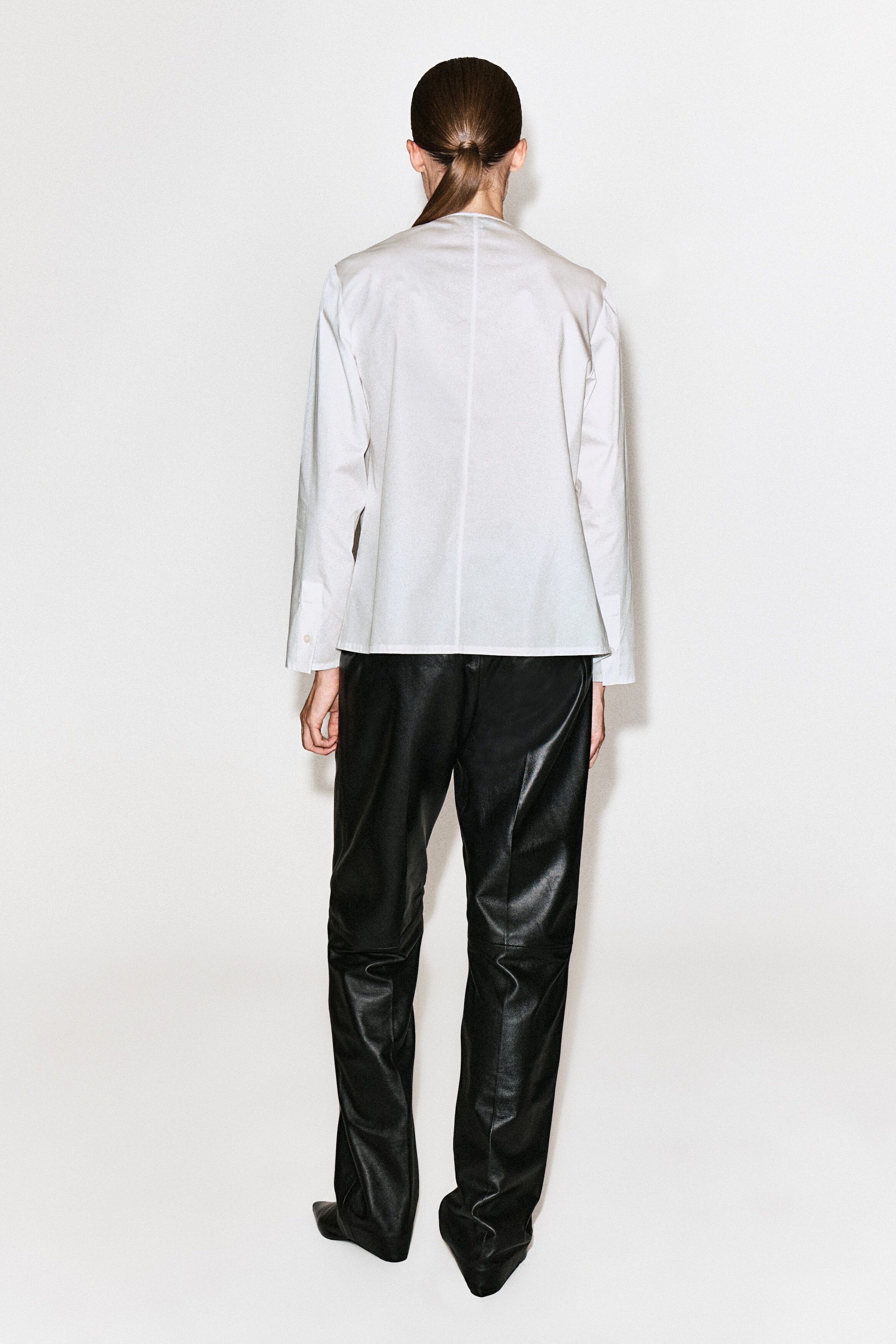 A white cotton blouse, featuring a center back seam and long sleeves with buttoned cuffs, is being worn by the model and styled with black leather trousers.
