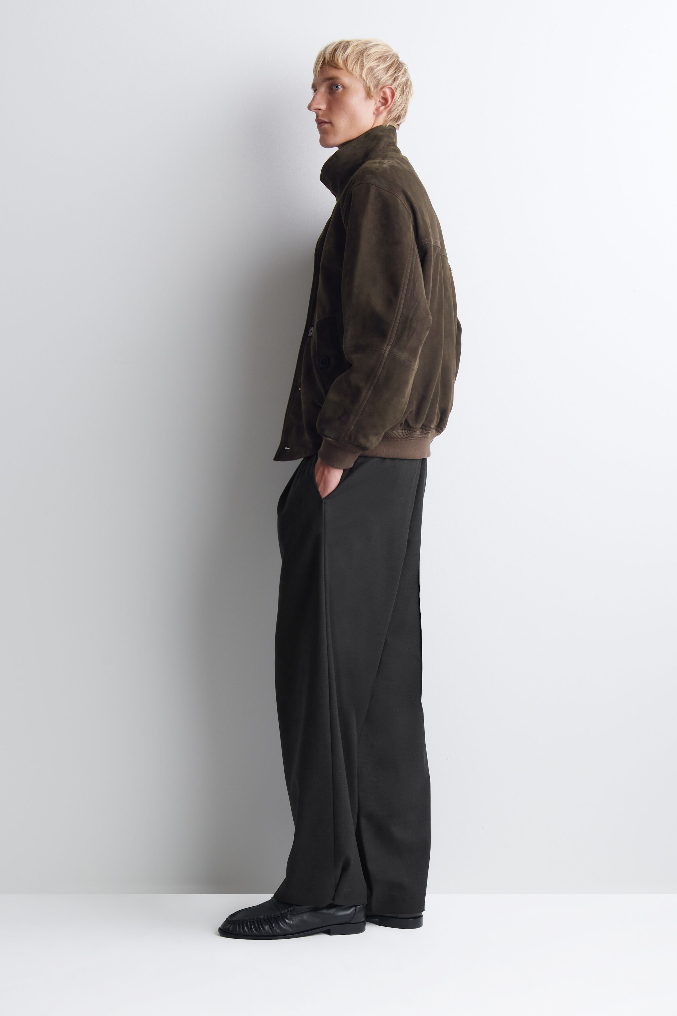 The wool relaxed-fit barrel-leg trousers drape in a curved, wide-leg silhouette, shown from the side profile facing left to highlight their structured shape. The trousers pool slightly over black leather shoes and feature a relaxed fit, with one hand resting in the side pocket. The wool relaxed-fit barrel-leg trousers drape in a curved, wide-leg silhouette, shown from the side profile facing left to highlight their structured shape. The trousers pool slightly over black leather shoes and feature a relaxed fit, with one hand resting in the side pocket.