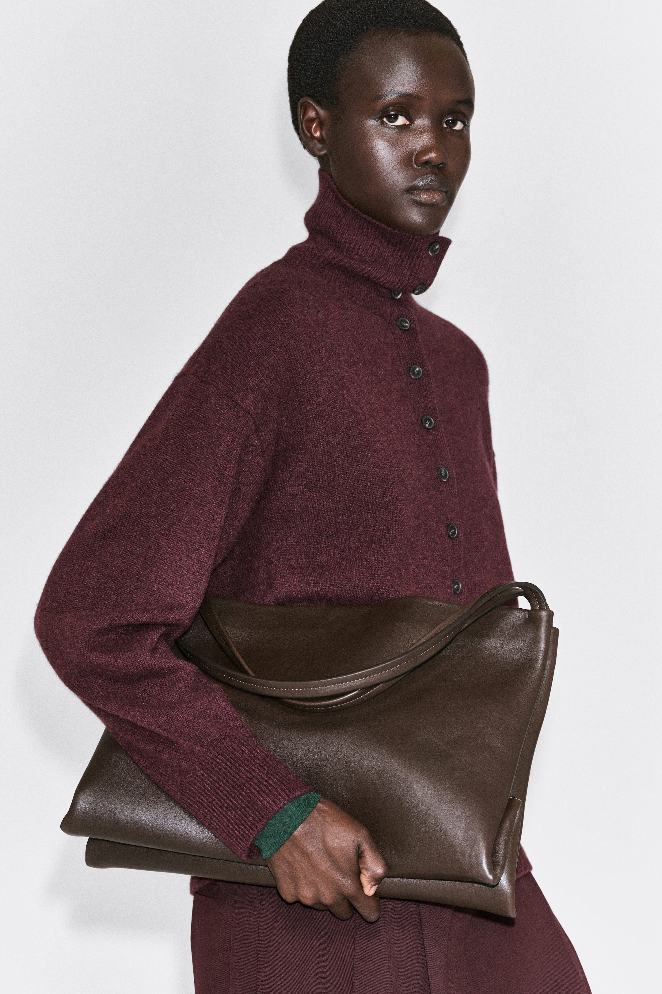 This burgundy cashmere cardigan, facing right, showcases a high buttoned funnel neck, dropped shoulders, and sculpted sleeves with long ribbed cuffs. The model is holding a large dark brown leather bag under their arm, highlighting the garment's relaxed silhouette and soft knit texture. This burgundy cashmere cardigan, facing right, showcases a high buttoned funnel neck, dropped shoulders, and sculpted sleeves with long ribbed cuffs. The model is holding a large dark brown leather bag under their arm, highlighting the garment's relaxed silhouette and soft knit texture.