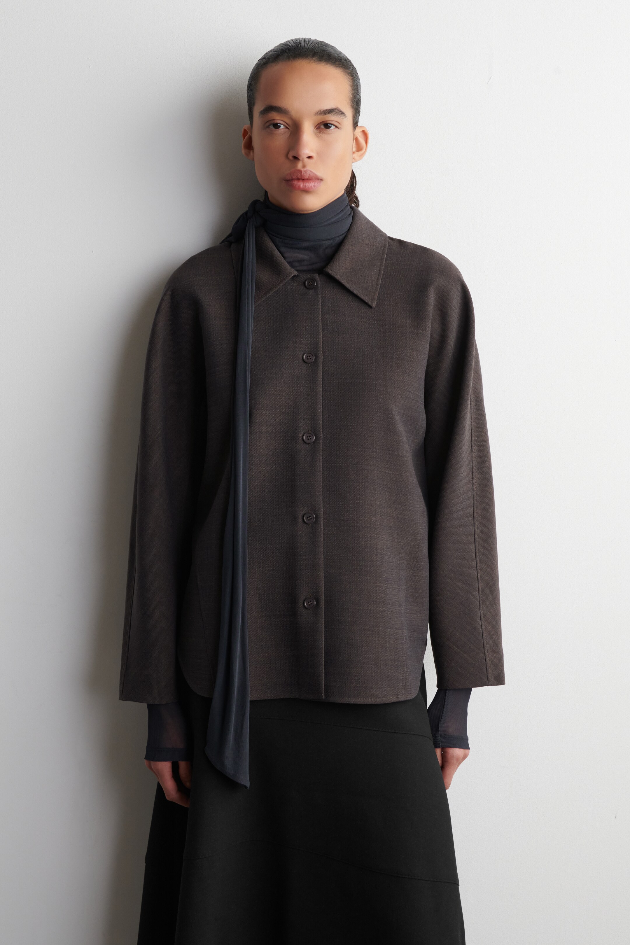 Größeres Bild ansehen: Eine dunkelbraune Jacke wird von vorne präsentiert, ihr klassischer Spitzkragen liegt über einem dunklen Rollkragenpullover. Sie verfügt über einen Verschluss mit vier Knöpfen und lange, gerade Ärmel, die eine entspannte, kastige Passform zeigen, die auf Hüfthöhe endet.