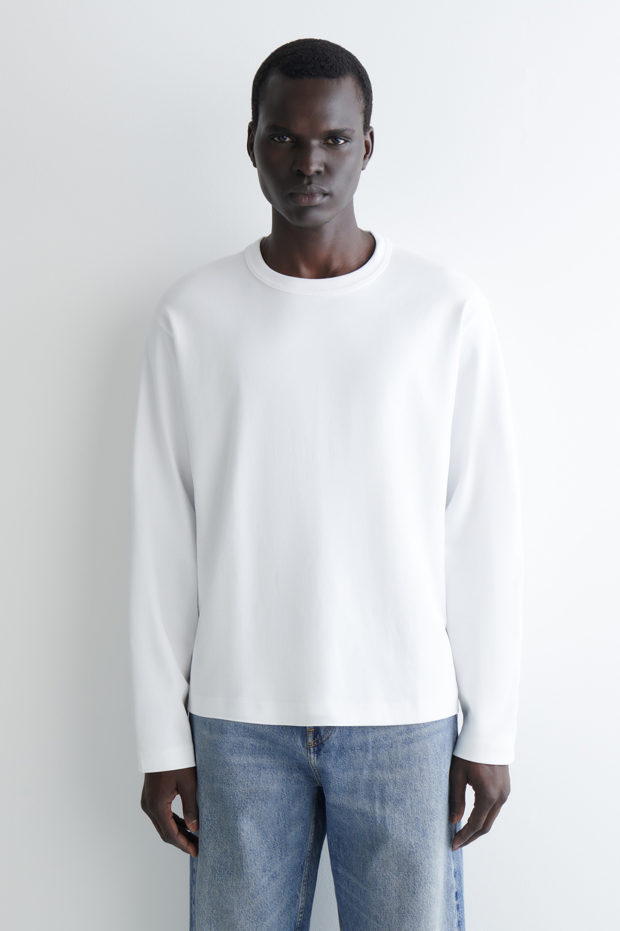 Agrandir l'image: Un t-shirt blanc à manches longues et col rond, doté d'une coupe fluide et décontractée avec des poignets simples et finis, est porté face avant avec son ourlet droit reposant sur un jean bleu.