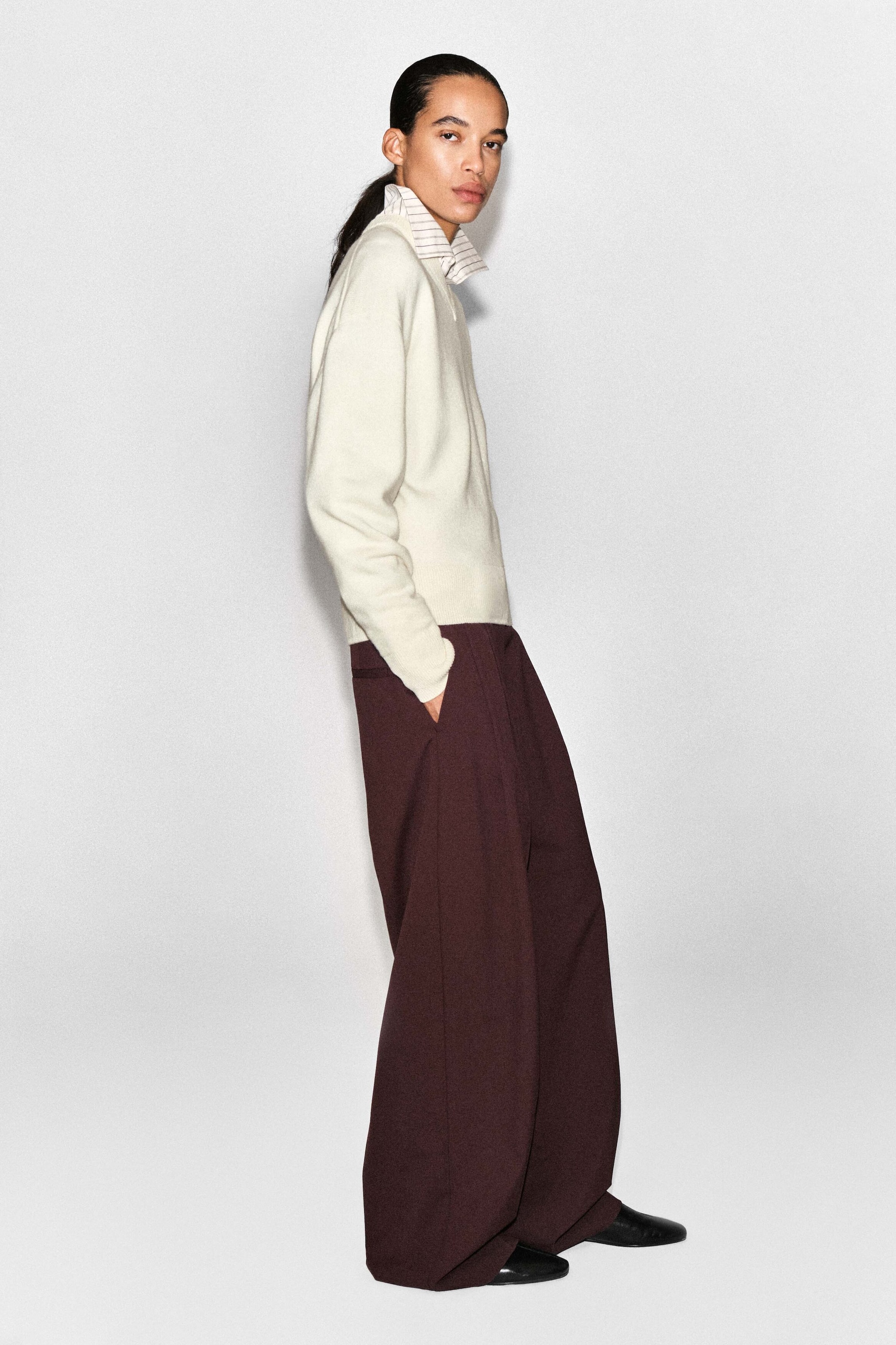 The merino wool layered V-neck jumper in cream is being worn by a model facing right, showcasing its relaxed fit and soft knit structure. The jumper features a V-neckline layered over a striped collared shirt, and displays ribbed cuffs and a ribbed hem. The merino wool layered V-neck jumper in cream is being worn by a model facing right, showcasing its relaxed fit and soft knit structure. The jumper features a V-neckline layered over a striped collared shirt, and displays ribbed cuffs and a ribbed hem.