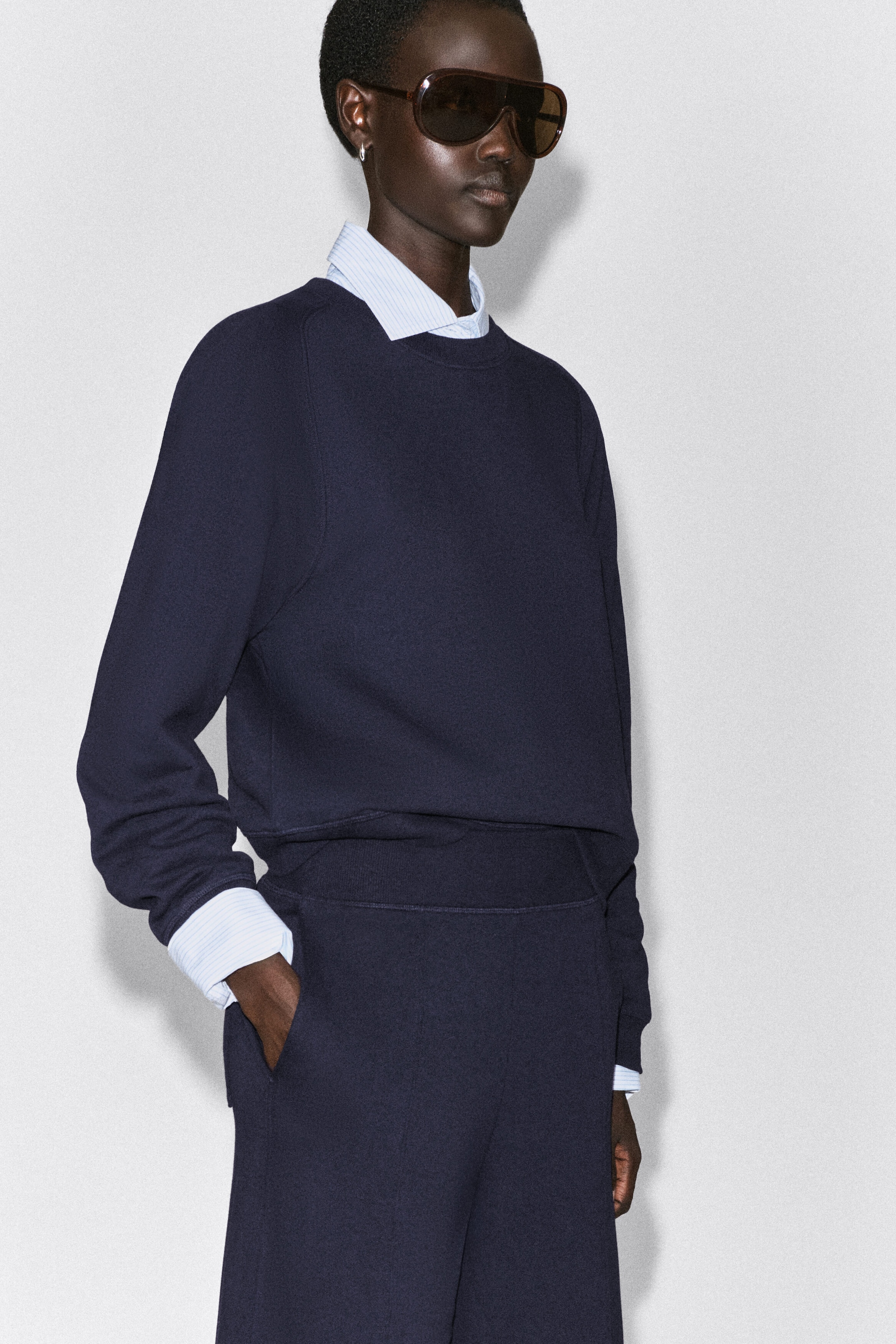 Die marineblauen Baumwoll-Jogger mit Barrel-Leg sind in einer High-Rise-Passform mit einem breiten, gerippten elastischen Bund und einer seitlichen Eingrifftasche zu sehen. Die Jogger werden mit einem passenden marineblauen Sweatshirt und einem darüber getragenen hellblau gestreiften Hemd kombiniert, wodurch ihr entspannter Fall und ihre bequeme Struktur betont werden. Die marineblauen Baumwoll-Jogger mit Barrel-Leg sind in einer High-Rise-Passform mit einem breiten, gerippten elastischen Bund und einer seitlichen Eingrifftasche zu sehen. Die Jogger werden mit einem passenden marineblauen Sweatshirt und einem darüber getragenen hellblau gestreiften Hemd kombiniert, wodurch ihr entspannter Fall und ihre bequeme Struktur betont werden.