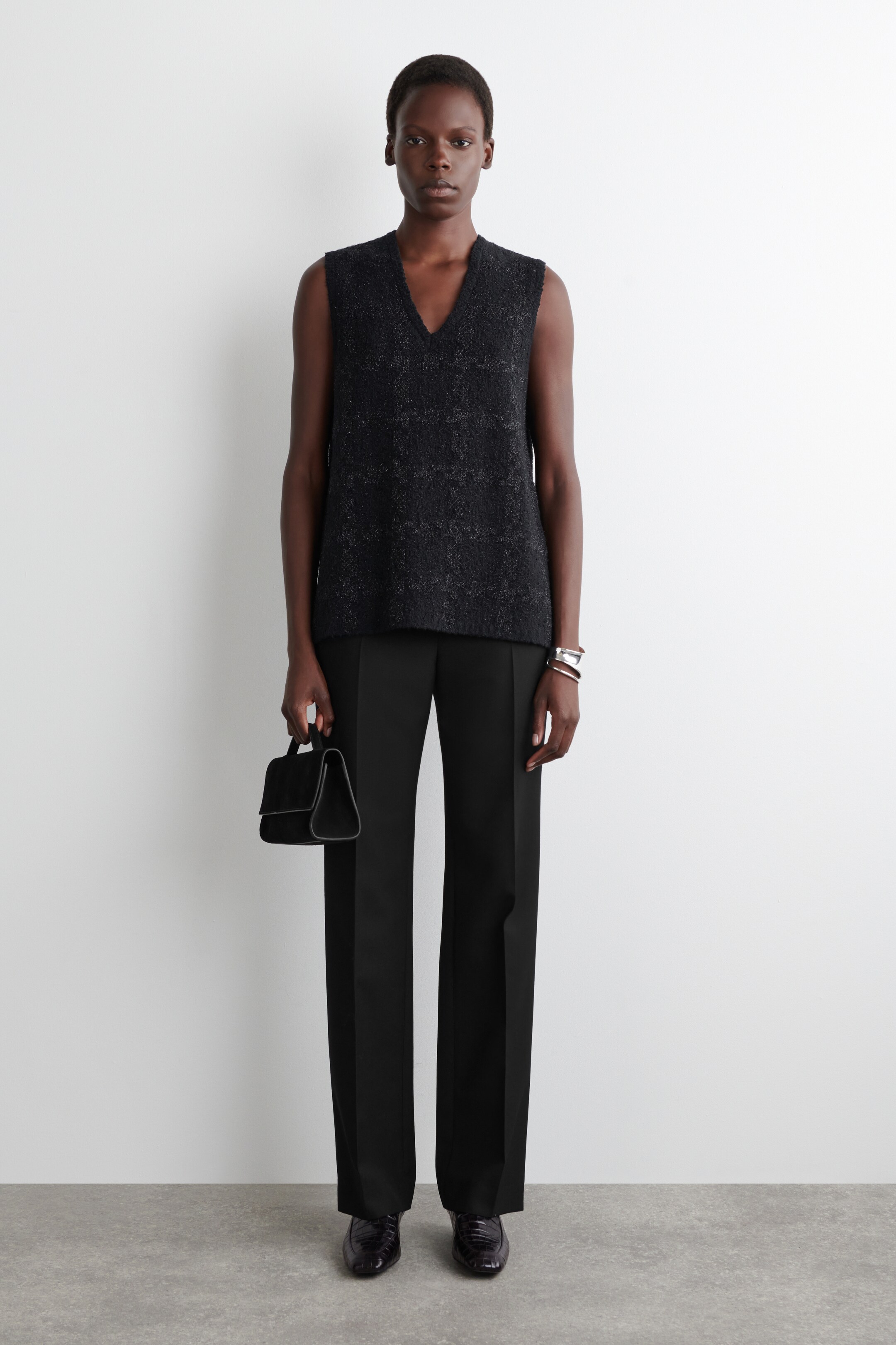 Agrandir l'image: Un haut noir sans manches avec un décolleté en V, doté d'une finition texturée, est présenté avec un pantalon noir à jambes droites en vue de face complète. Des mocassins noirs brillants sont portés, et un petit sac à main structuré noir est porté.