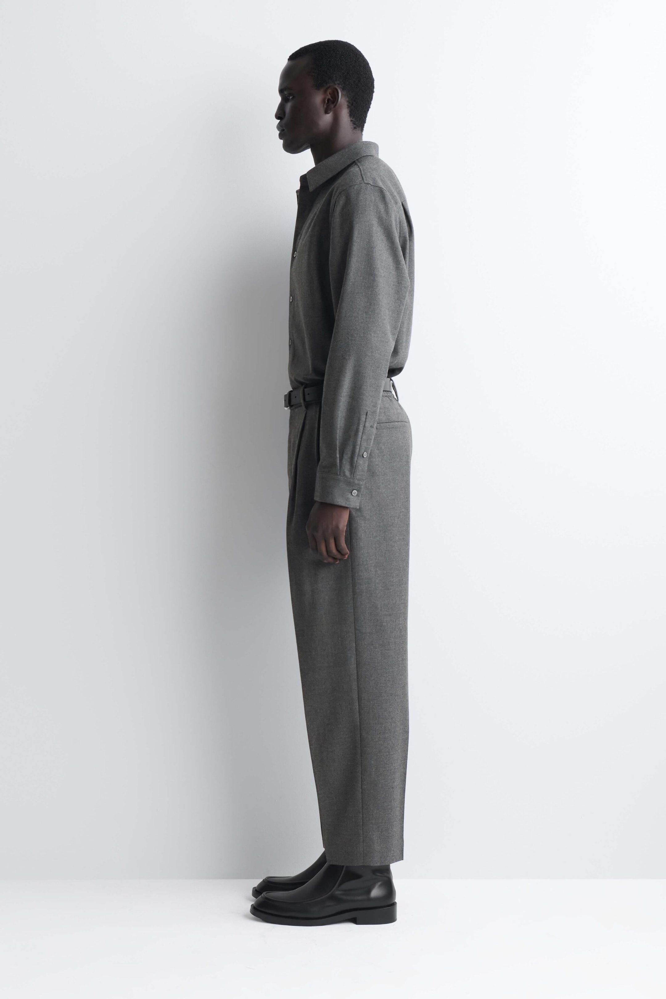 Ein passendes Set aus grauem Langarmhemd und Hose wird in linker Seitenansicht gezeigt. Das Hemd mit Knopfleiste ist in eine strukturierte Hose mit geradem Bein gesteckt, die mit einem schmalen schwarzen Gürtel und schwarzen Lederstiefeln kombiniert wird. Ein passendes Set aus grauem Langarmhemd und Hose wird in linker Seitenansicht gezeigt. Das Hemd mit Knopfleiste ist in eine strukturierte Hose mit geradem Bein gesteckt, die mit einem schmalen schwarzen Gürtel und schwarzen Lederstiefeln kombiniert wird.