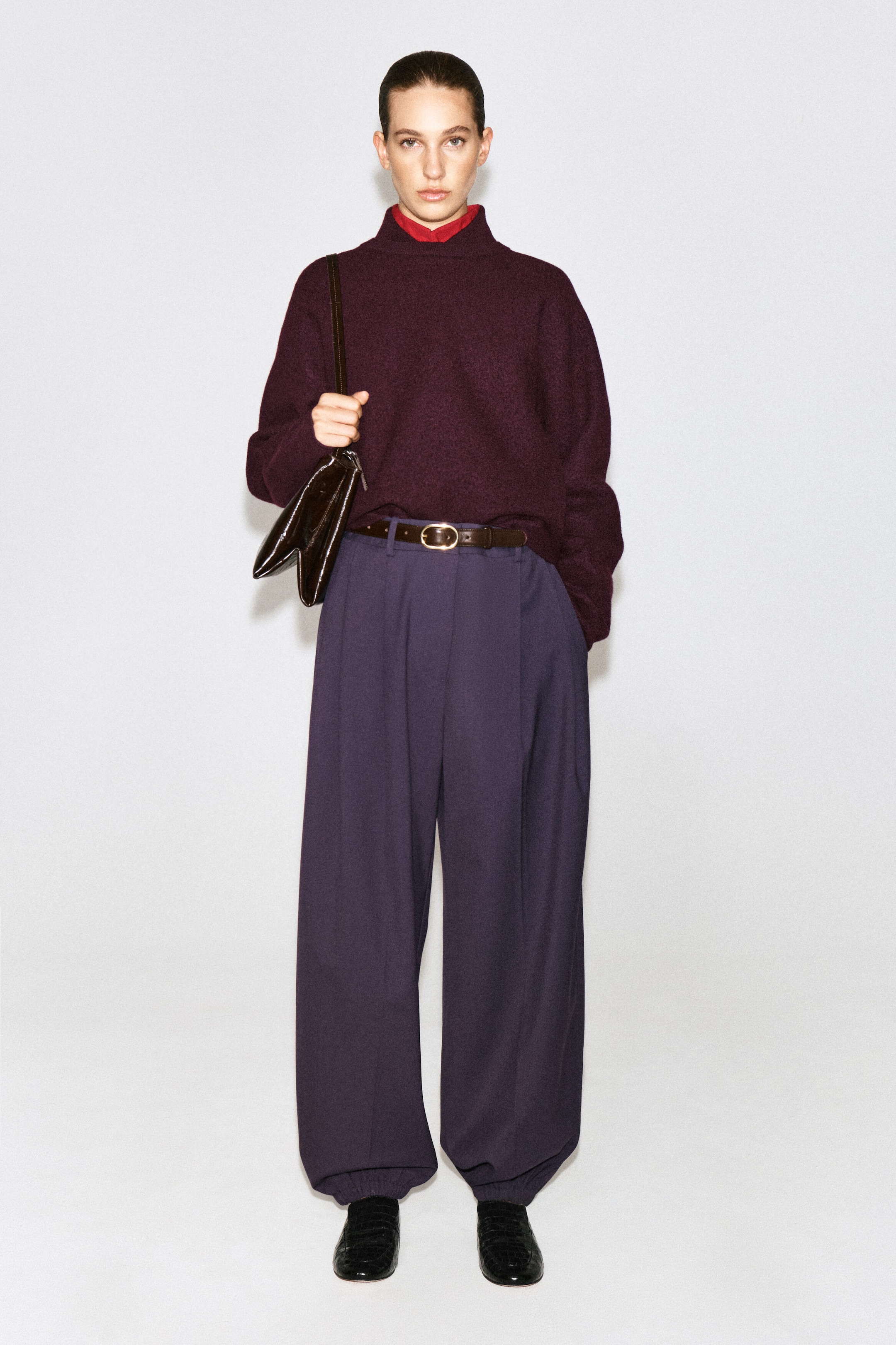 Un chandail en laine bouillie à col superposé, couleur bordeaux profond, est porté par le mannequin. Il présente un col cheminée haut qui laisse apparaître un col rouge éclatant en dessous. Le chandail à coupe décontractée est rentré dans un pantalon mauve à jambes larges et ceinturé, et il est agencé avec un sac à bandoulière foncé tenu d’un côté. Un chandail en laine bouillie à col superposé, couleur bordeaux profond, est porté par le mannequin. Il présente un col cheminée haut qui laisse apparaître un col rouge éclatant en dessous. Le chandail à coupe décontractée est rentré dans un pantalon mauve à jambes larges et ceinturé, et il est agencé avec un sac à bandoulière foncé tenu d’un côté.