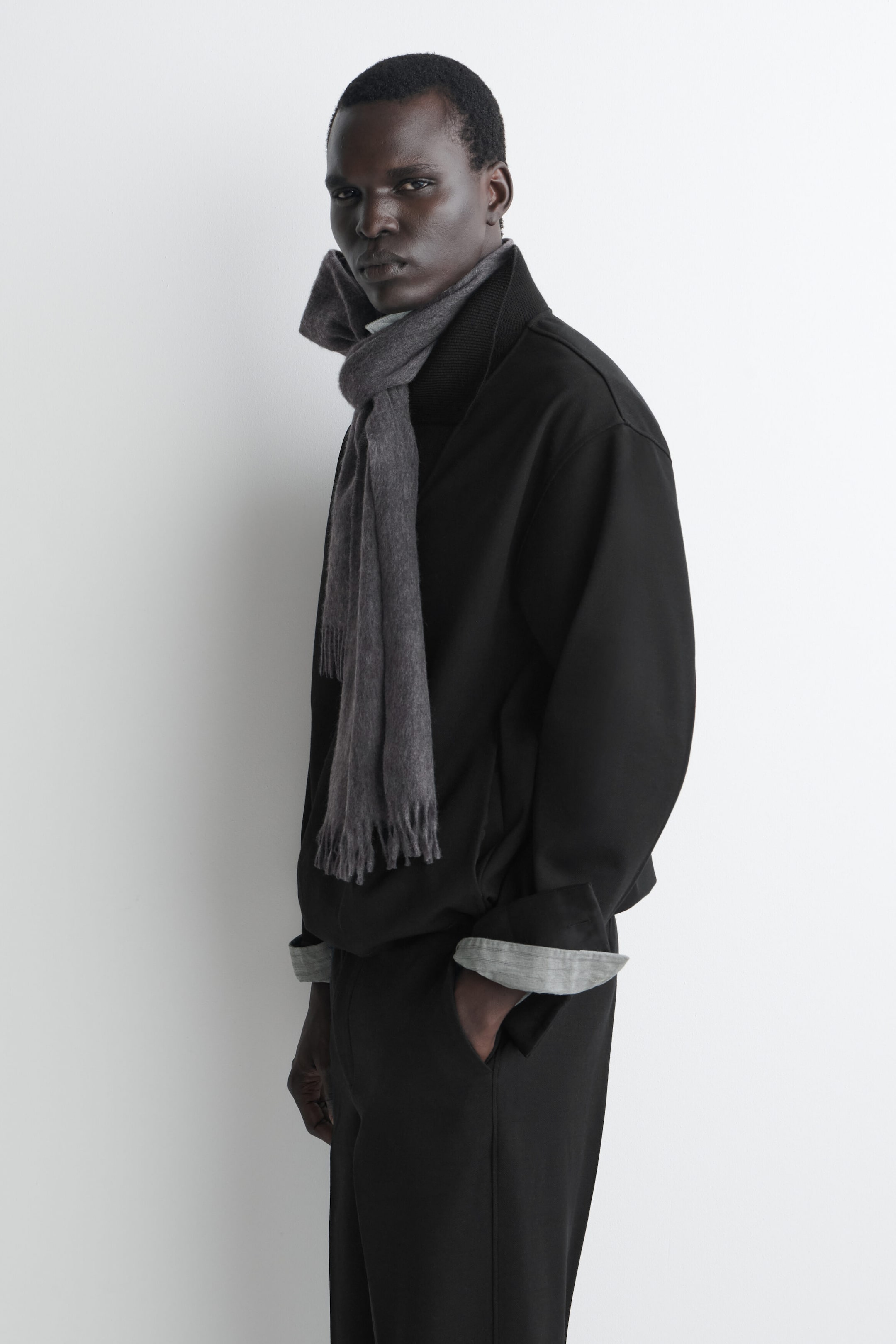 A grey cashmere scarf with fringed ends is worn knotted around the neck, with its draped ends hanging down the front of a black jacket. The scarf is shown on a model facing three-quarters left, highlighting its soft texture and classic design. A grey cashmere scarf with fringed ends is worn knotted around the neck, with its draped ends hanging down the front of a black jacket. The scarf is shown on a model facing three-quarters left, highlighting its soft texture and classic design.