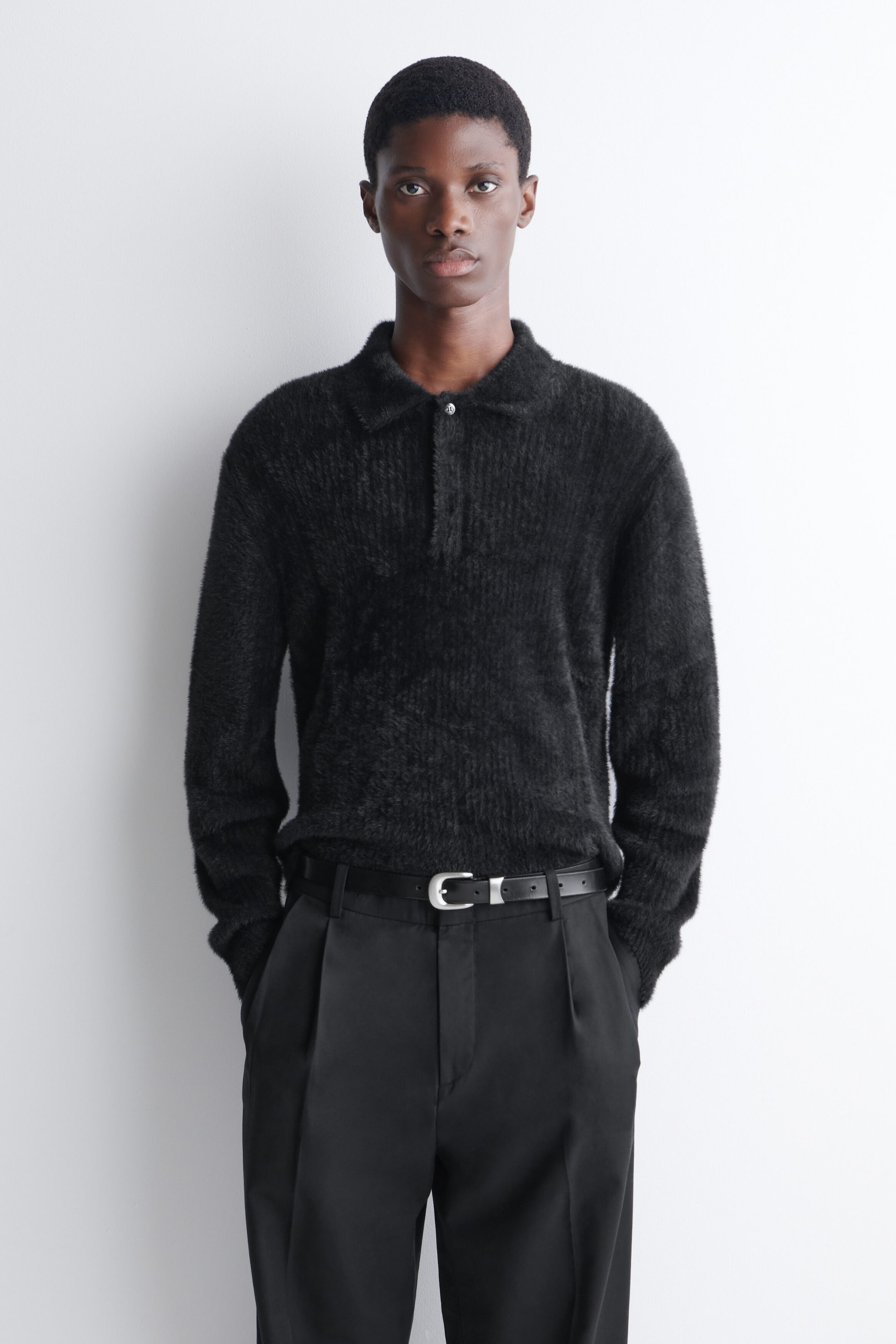 A black, brushed-knit long-sleeve polo shirt with a fuzzy texture and a classic collar is worn by the model. The shirt is tucked into black tailored trousers secured with a silver-buckle belt. A black, brushed-knit long-sleeve polo shirt with a fuzzy texture and a classic collar is worn by the model. The shirt is tucked into black tailored trousers secured with a silver-buckle belt.