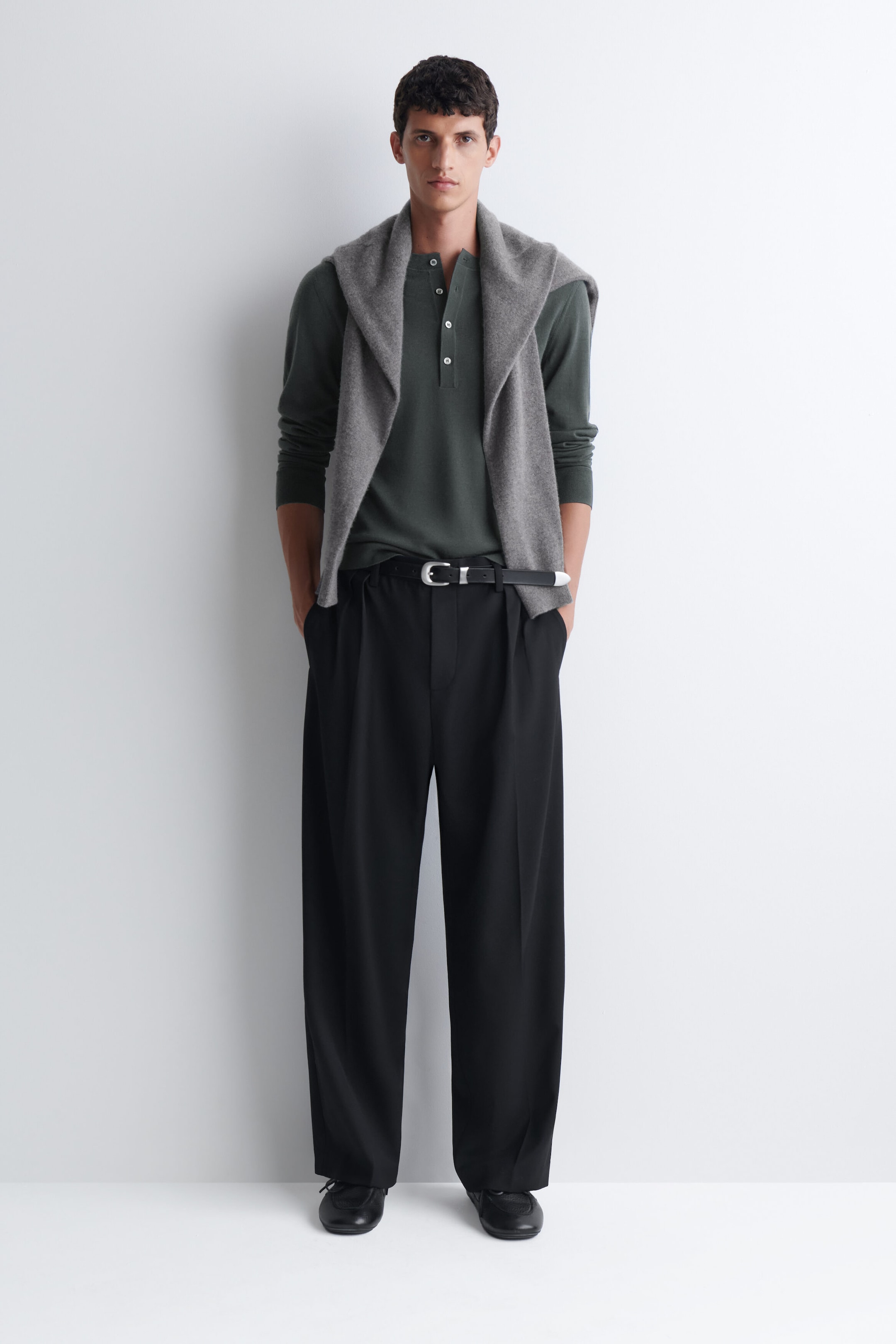 The slim merino wool henley top in dark green is worn by the model, featuring a button placket and long sleeves. The top is tucked into pleated black trousers with a belt and styled with a grey knit sweater draped over the shoulders.