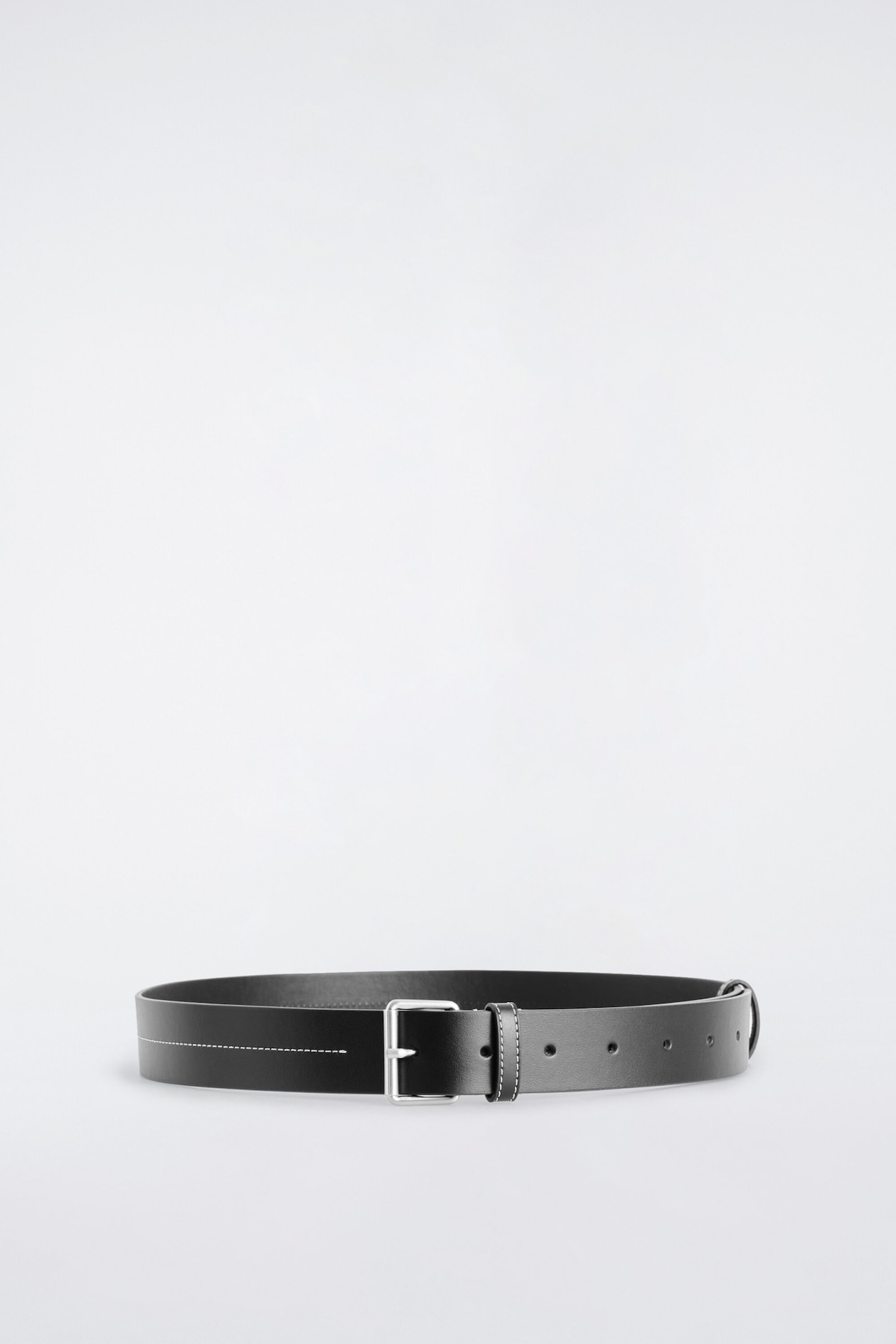 A black leather belt is resting in a fastened loop facing forward, showcasing a rectangular silver-tone buckle and a matching leather keeper with white contrast stitching. A horizontal line of white contrast stitching runs along the left side of the smooth strap, which features several adjustment holes on the right.