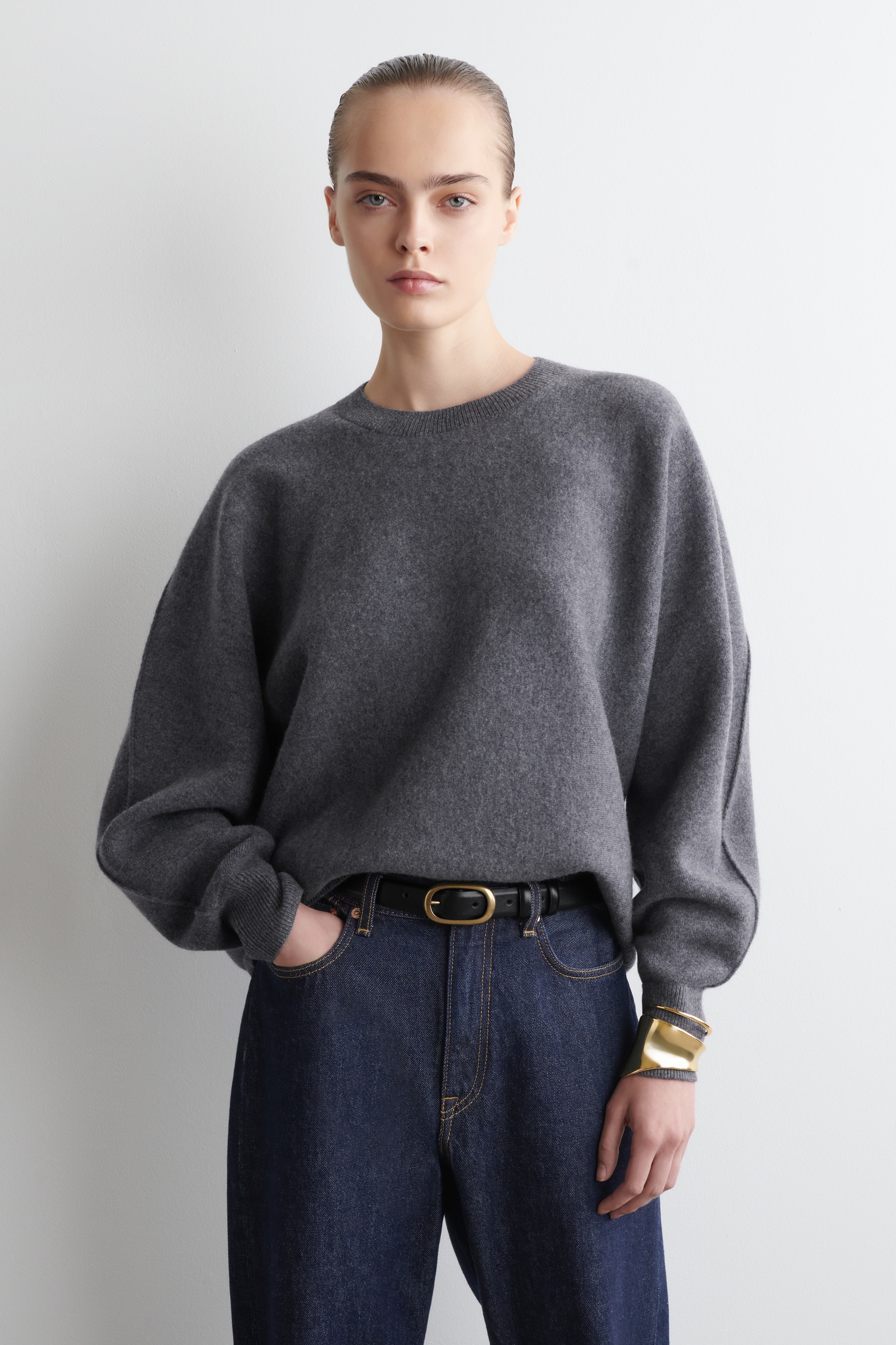 Grösseres Bild ansehen: Ein dunkelgrauer Pullover mit Rundhalsausschnitt wird frontseitig getragen und zeigt seine lockere Passform, überschnittene Schultern und ein sichtbares Nahtdetail, das entlang des äußeren Arms verläuft. Der Pullover hat gerippte Bündchen und wird mit dunkel gewaschenen Jeans, einem schwarzen Gürtel mit goldener Schnalle und einem goldenen Armreif, der über dem Ärmel getragen wird, gestylt.
