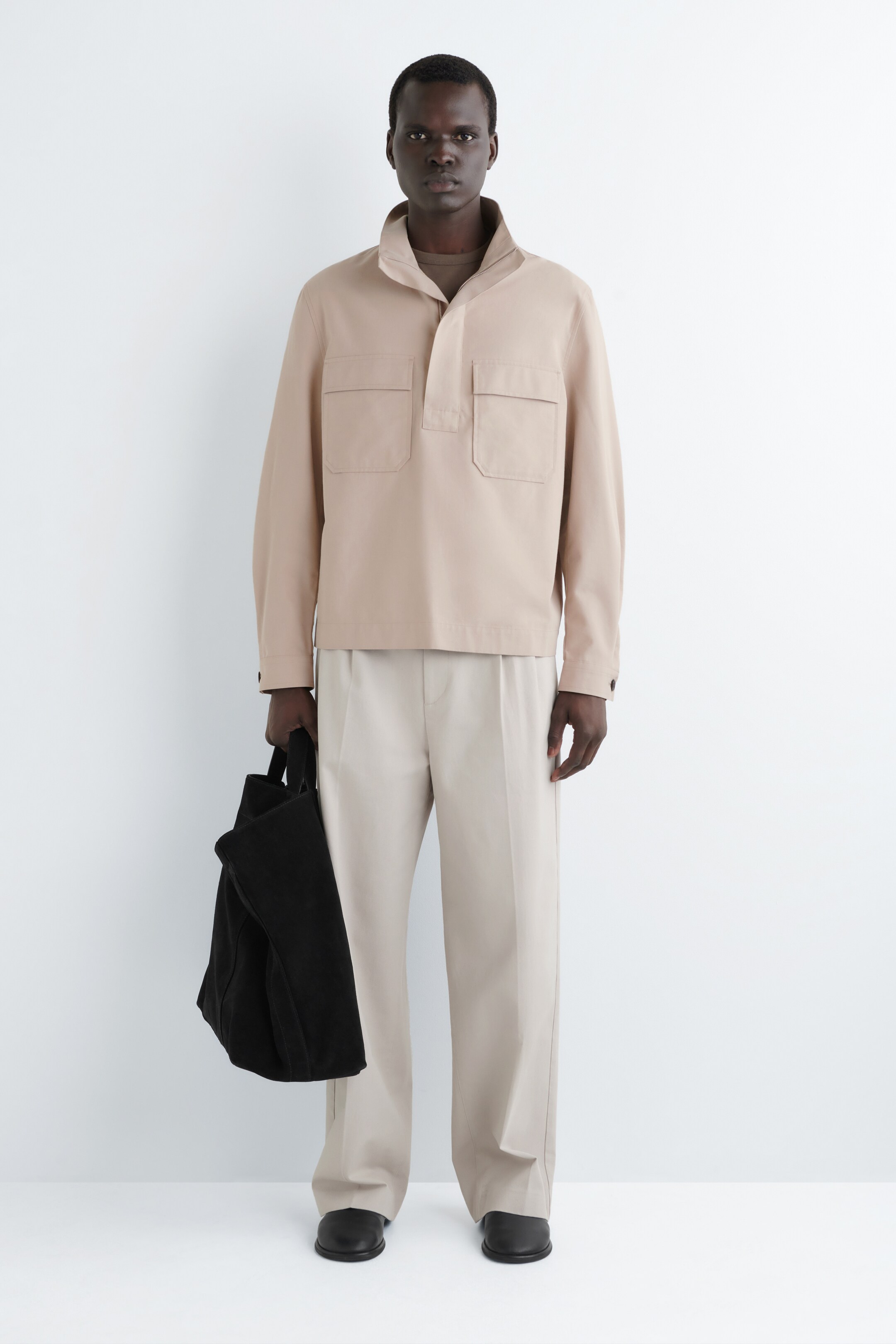 Ver imagen más grande: Un pulóver de manga larga color beige se presenta de frente, cuenta con un cuello stand, una tapeta parcial y dos bolsillos con solapa en el pecho. Está superpuesto sobre una camiseta marrón con cuello redondo y se combina con pantalones anchos con pliegues en beige claro, zapatos negros sin cordones y un bolso tote negro.