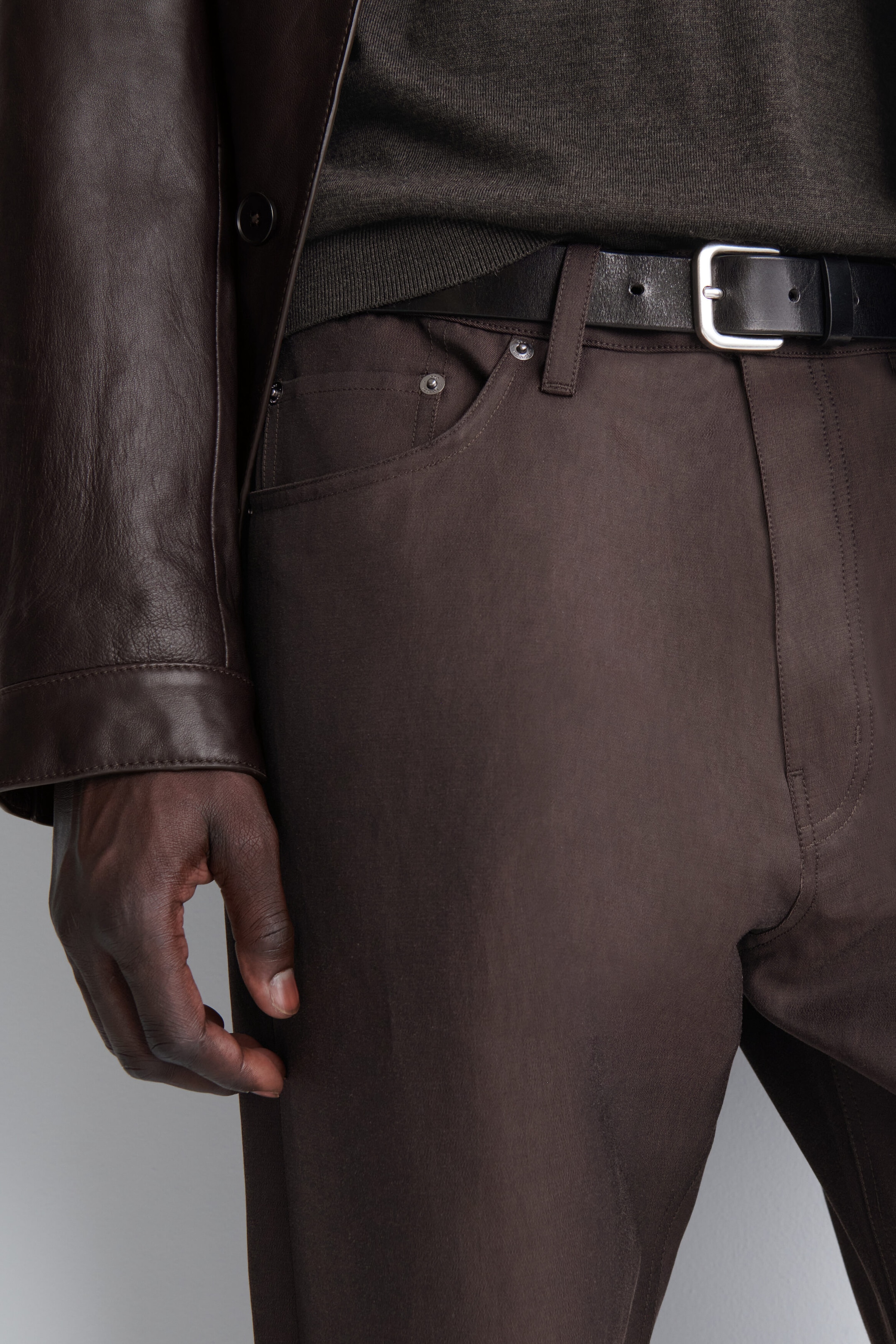 The signature regular-fit straight-leg jeans are shown from the front in a close-up view, highlighting their dark brown fabric, tonal stitching, and classic five-pocket styling with metal rivets. The jeans are being worn with a black leather belt threaded through the belt loops, a dark grey knit jumper, and a dark brown leather jacket. The signature regular-fit straight-leg jeans are shown from the front in a close-up view, highlighting their dark brown fabric, tonal stitching, and classic five-pocket styling with metal rivets. The jeans are being worn with a black leather belt threaded through the belt loops, a dark grey knit jumper, and a dark brown leather jacket.