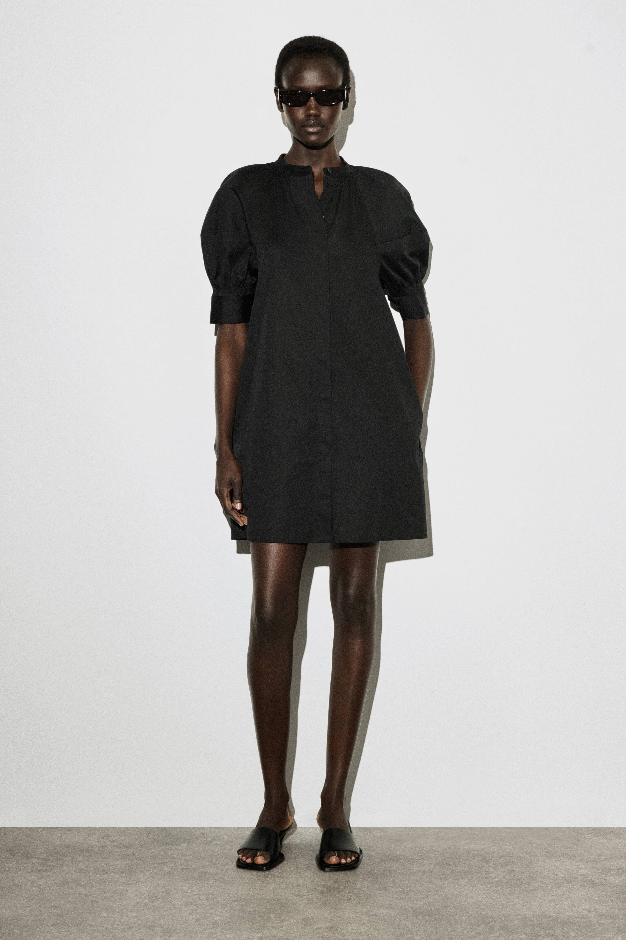 Agrandir l'image: La robe noire courte présente un col montant avec une ouverture en V, des manches bouffantes longueur coude avec poignets, et une silhouette trapèze décontractée. Elle est conçue avec des poches latérales visibles.