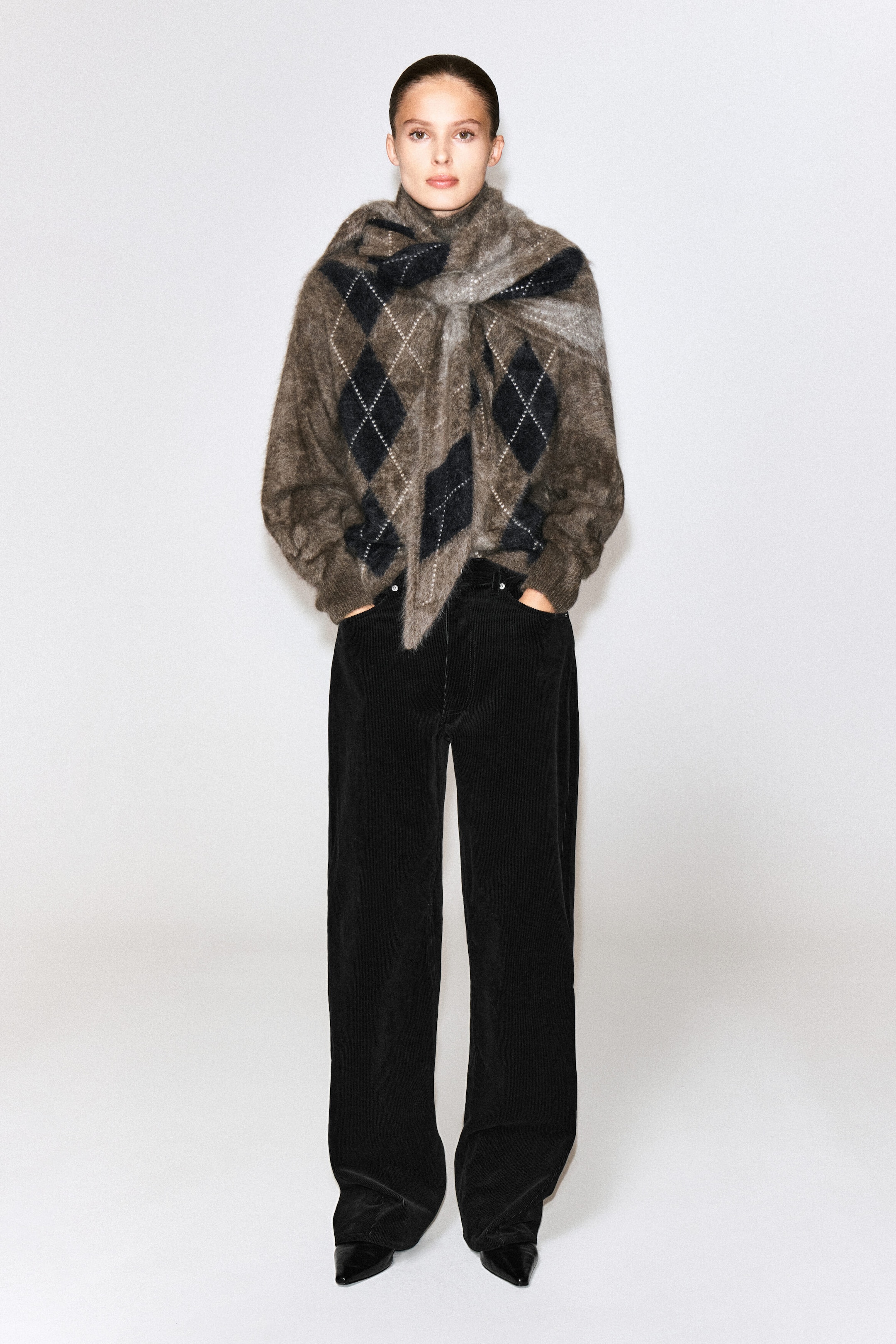 A brushed cashmere argyle triangle scarf in shades of brown, grey, and black is worn by the model, draped over the shoulders and loosely knotted at the front. The textured scarf is styled over a matching long-sleeved argyle knit sweater and paired with black trousers. A brushed cashmere argyle triangle scarf in shades of brown, grey, and black is worn by the model, draped over the shoulders and loosely knotted at the front. The textured scarf is styled over a matching long-sleeved argyle knit sweater and paired with black trousers.