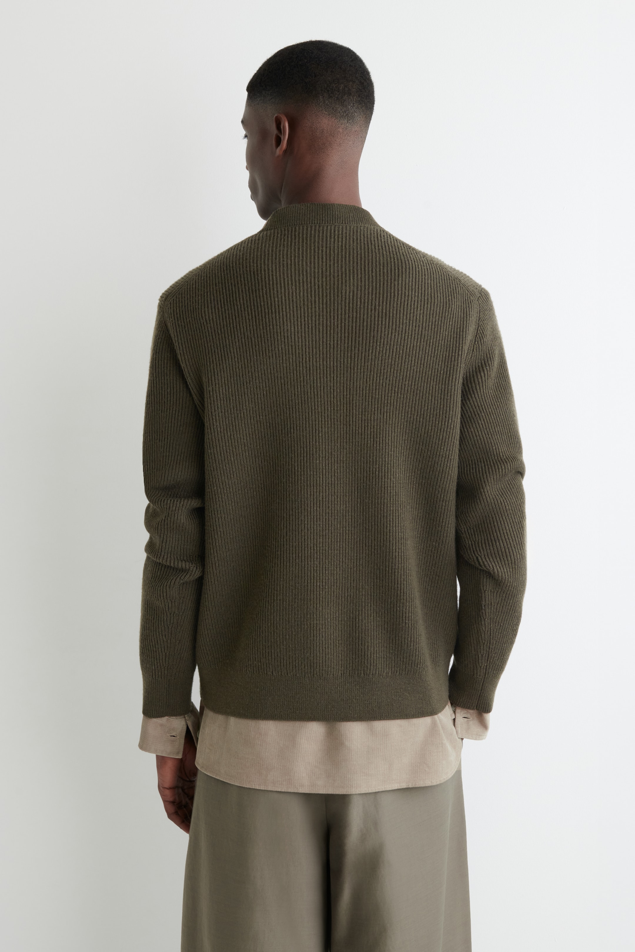 Grösseres Bild ansehen: Ein olivgrüner gerippter Strickpullover wird getragen, von hinten gesehen, mit einem niedrigen Stehkragen, langen Ärmeln und einer vertikalen Rippstruktur. Ein helles Hemd wird darunter getragen, dessen Saum und Manschetten unter dem Pullover sichtbar sind.