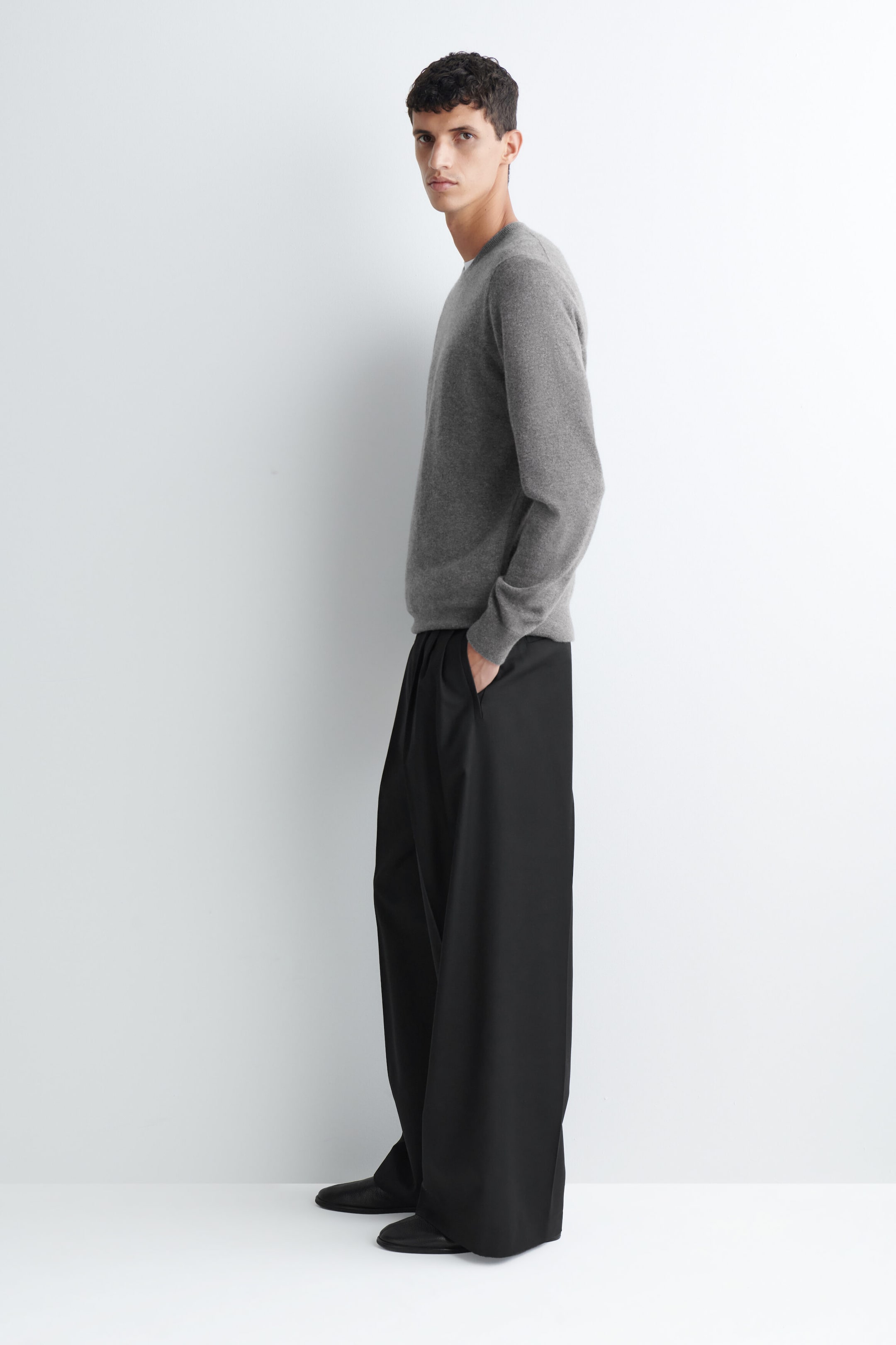 Die Model trägt plissierte, locker geschnittene schwarze Wide-Leg-Hose und steht im Profil, wobei eine Hand in der Seitentasche ruht. Die Hose ist mit einem grauen langärmeligen Rundhalspullover und schwarzen Schuhen kombiniert.