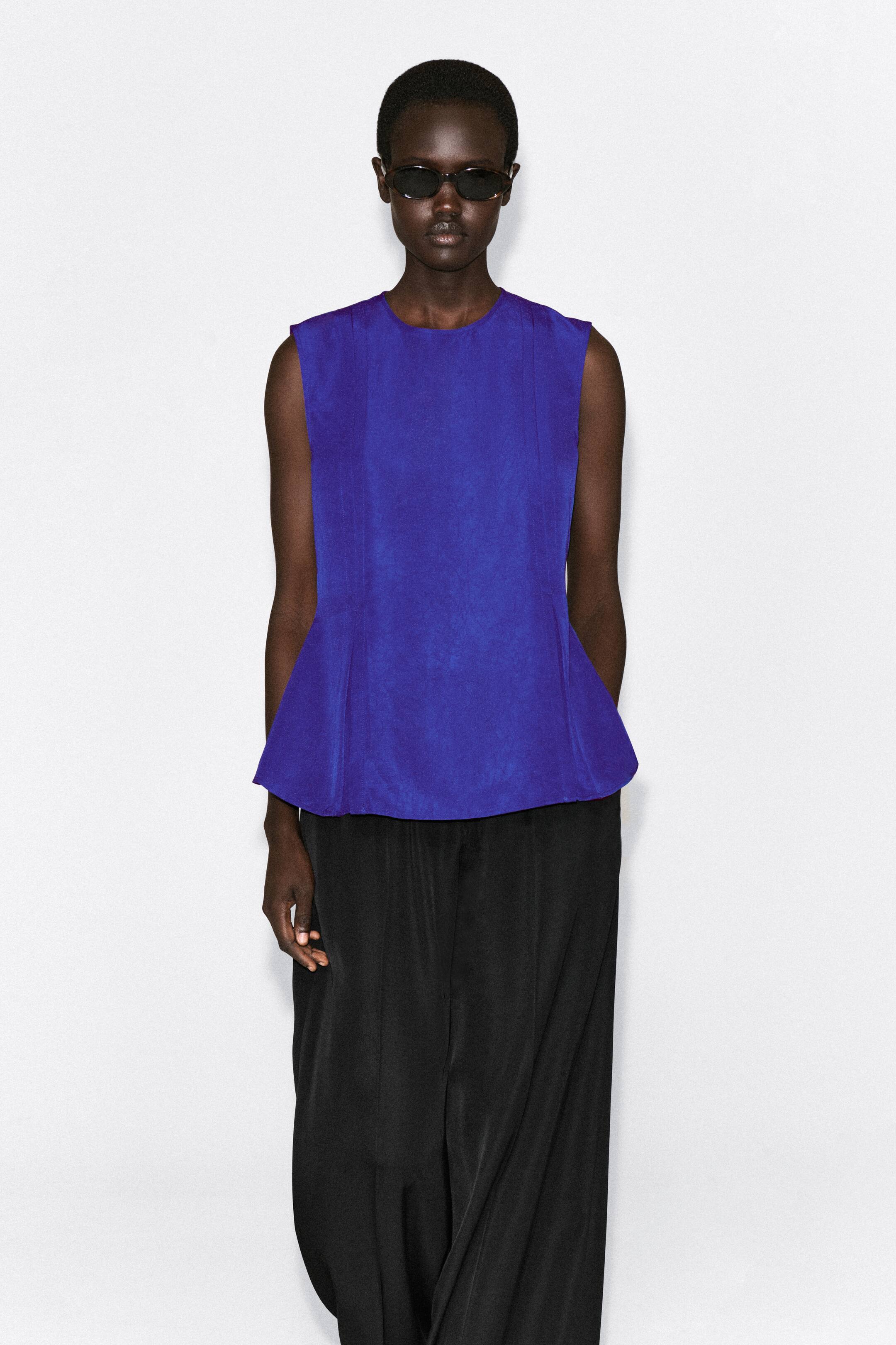 Visualizza immagine più grande: Un vivace top peplum blu senza maniche è mostrato frontalmente, con girocollo e pieghe verticali strutturate che si aprono sull’orlo. Il top è abbinato a pantaloni neri dalla vestibilità rilassata, mettendo in risalto la sua silhouette svasata.