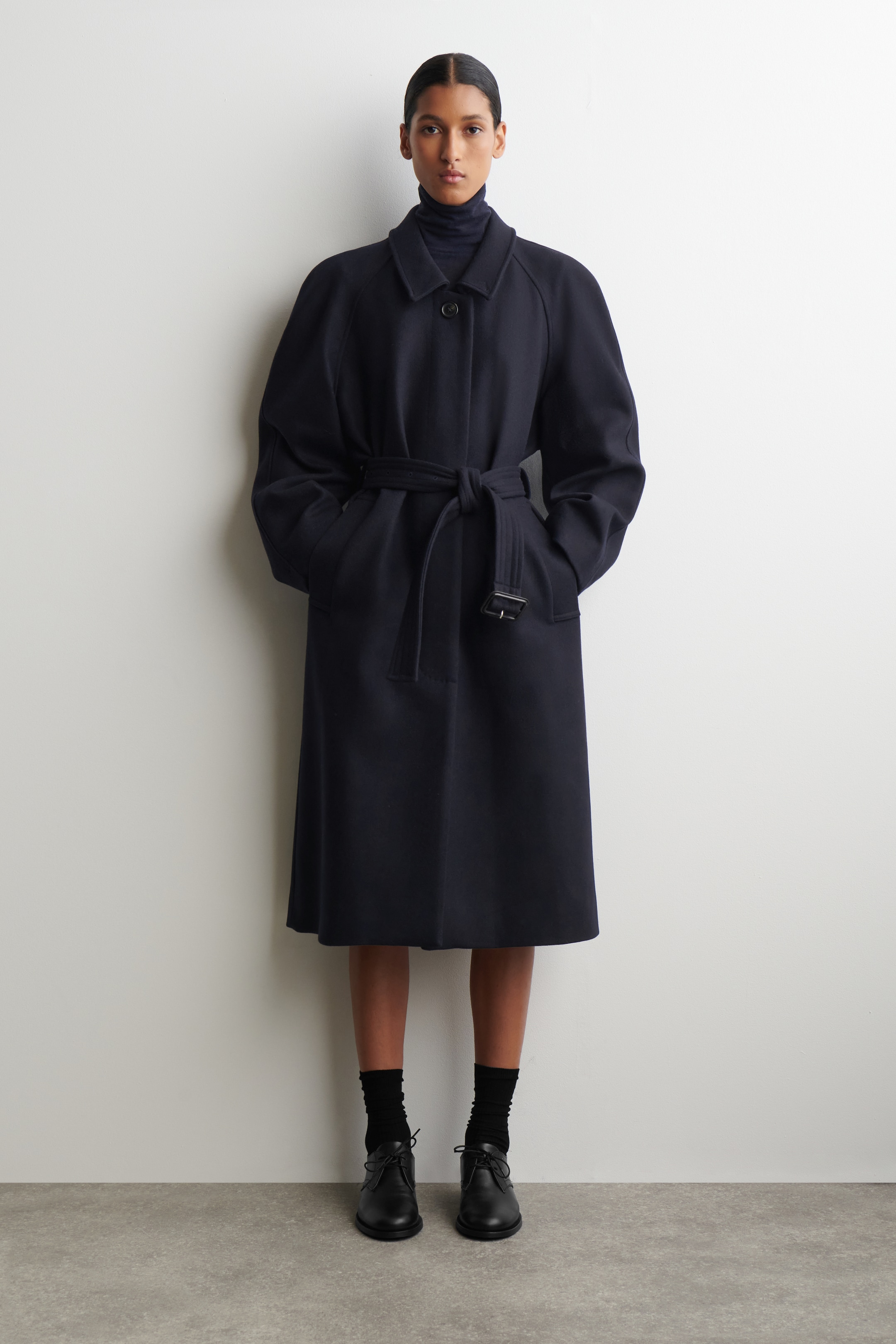 Agrandir l'image: Un manteau de couleur sombre est présenté en vue de face, avec un col pointu classique et des manches raglan volumineuses. Il est ceinturé à la taille par une ceinture en tissu assorti avec une boucle rectangulaire, et tombe à une longueur midi sur un col roulé foncé.