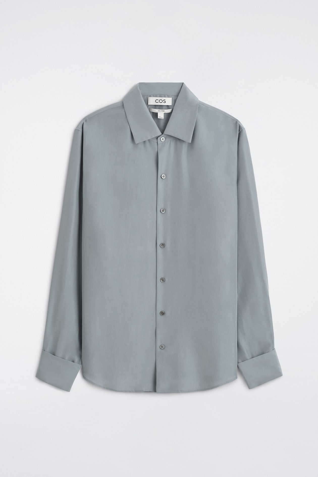 A relaxed silk shirt in a muted grey-blue shade is presented from the front, showing a classic point collar and a central button-down placket. The long sleeves feature wide, folded cuffs, and the design finishes with a subtly curved hem.
