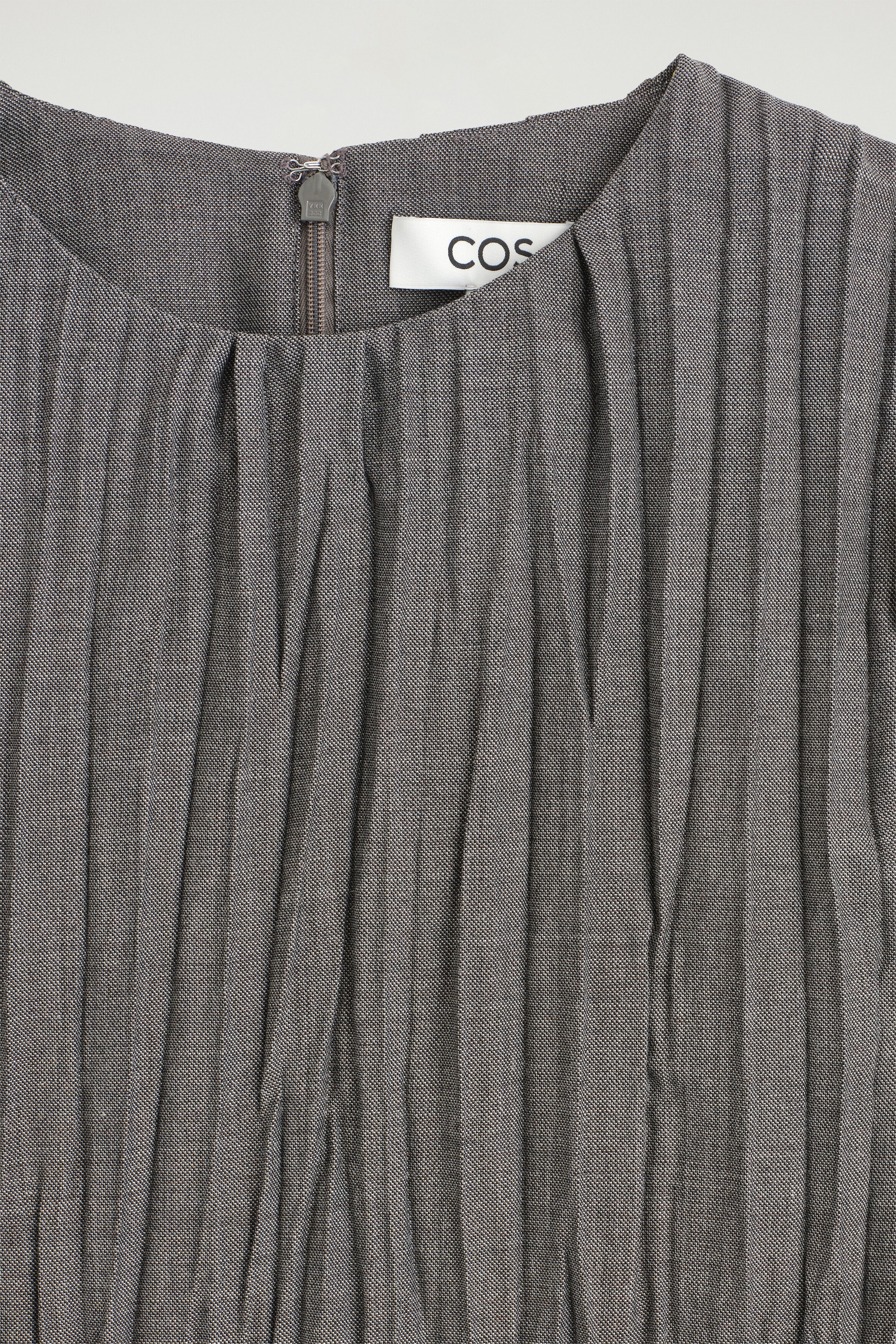 A gray textured top is shown in close-up, highlighting its round neckline with vertical pleats that drape downward. The garment features a short zipper closure with a hook-and-eye fastener at the center neckline, and a white COS brand label is visible on the inside.