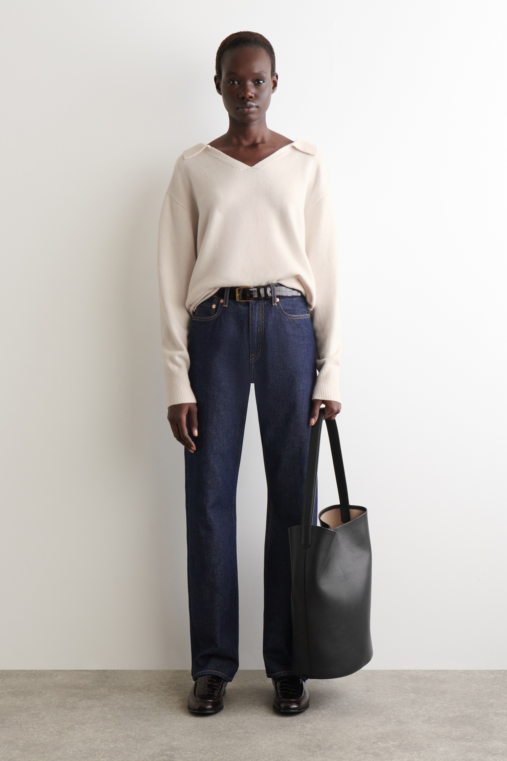 Visa större bild: En ljus krämfärgad V-ringad tröja, med en bred, öppen krage designad för att vila på axlarna, presenteras. Den är stylad med mörktvättade raka jeans, ett svart bälte och mörkbruna texturerade loafers, medan en svart ostrukturerad tote-väska hålls vid sidan.