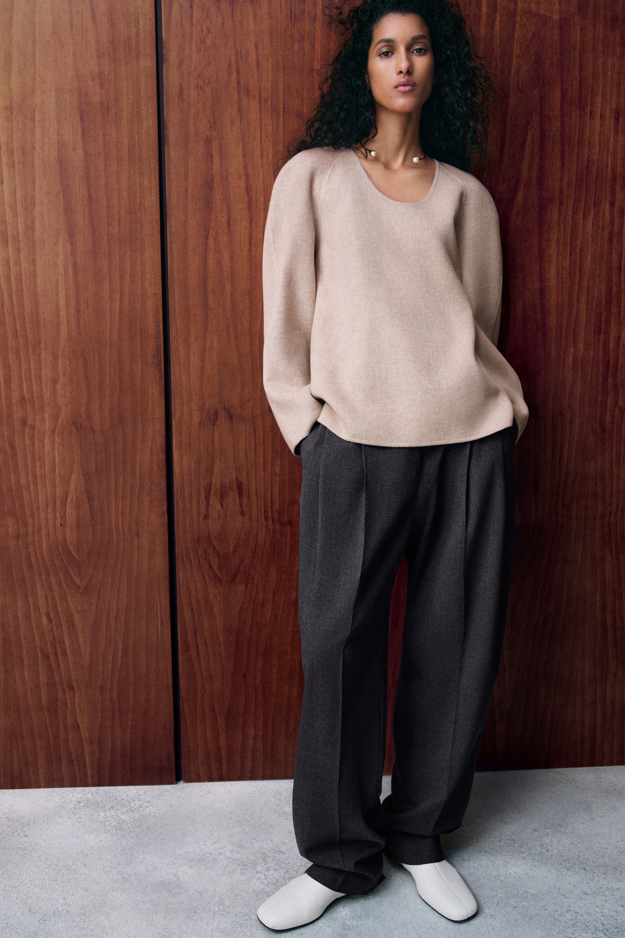 Größeres Bild anzeigen: Das beigefarbene DOUBLE-FACED WOOL TOP zeichnet sich durch einen Rundhalsausschnitt und lange Raglanärmel aus und bietet eine lockere Passform. Es wird mit dunkelgrauen Hosen aus einer Merino-Wollmischung getragen, die auffällige Frontfalten und eine Barrel-Leg-Silhouette aufweisen, von VORNE gesehen.