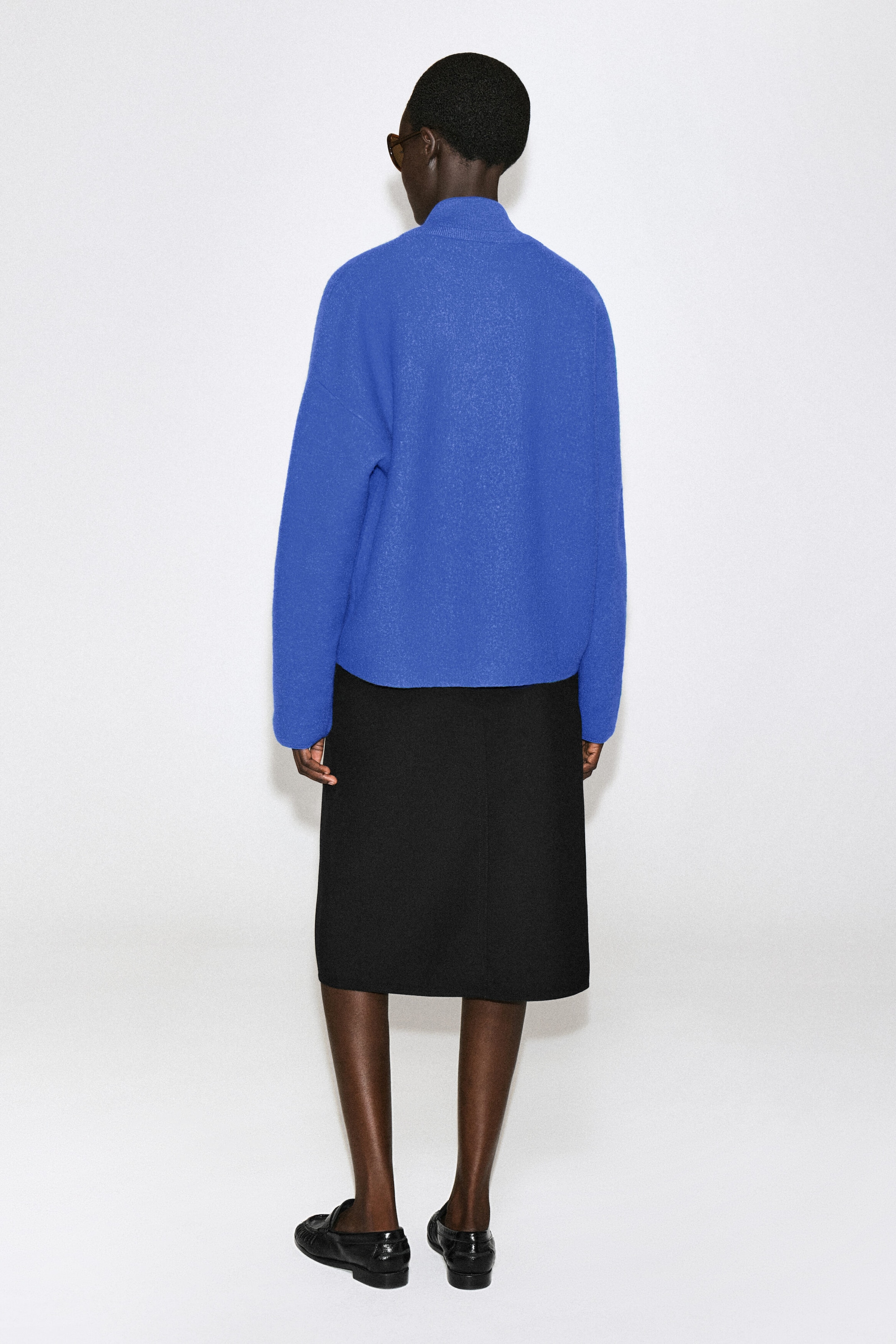 Le chandail en laine bouillie bleu vif, vu de dos, met en valeur une silhouette ample avec épaules tombantes, manches longues et col montant. Il tombe droit jusqu’aux hanches, porté par-dessus une jupe noire longueur genou pour souligner la netteté de son ourlet. Le chandail en laine bouillie bleu vif, vu de dos, met en valeur une silhouette ample avec épaules tombantes, manches longues et col montant. Il tombe droit jusqu’aux hanches, porté par-dessus une jupe noire longueur genou pour souligner la netteté de son ourlet.