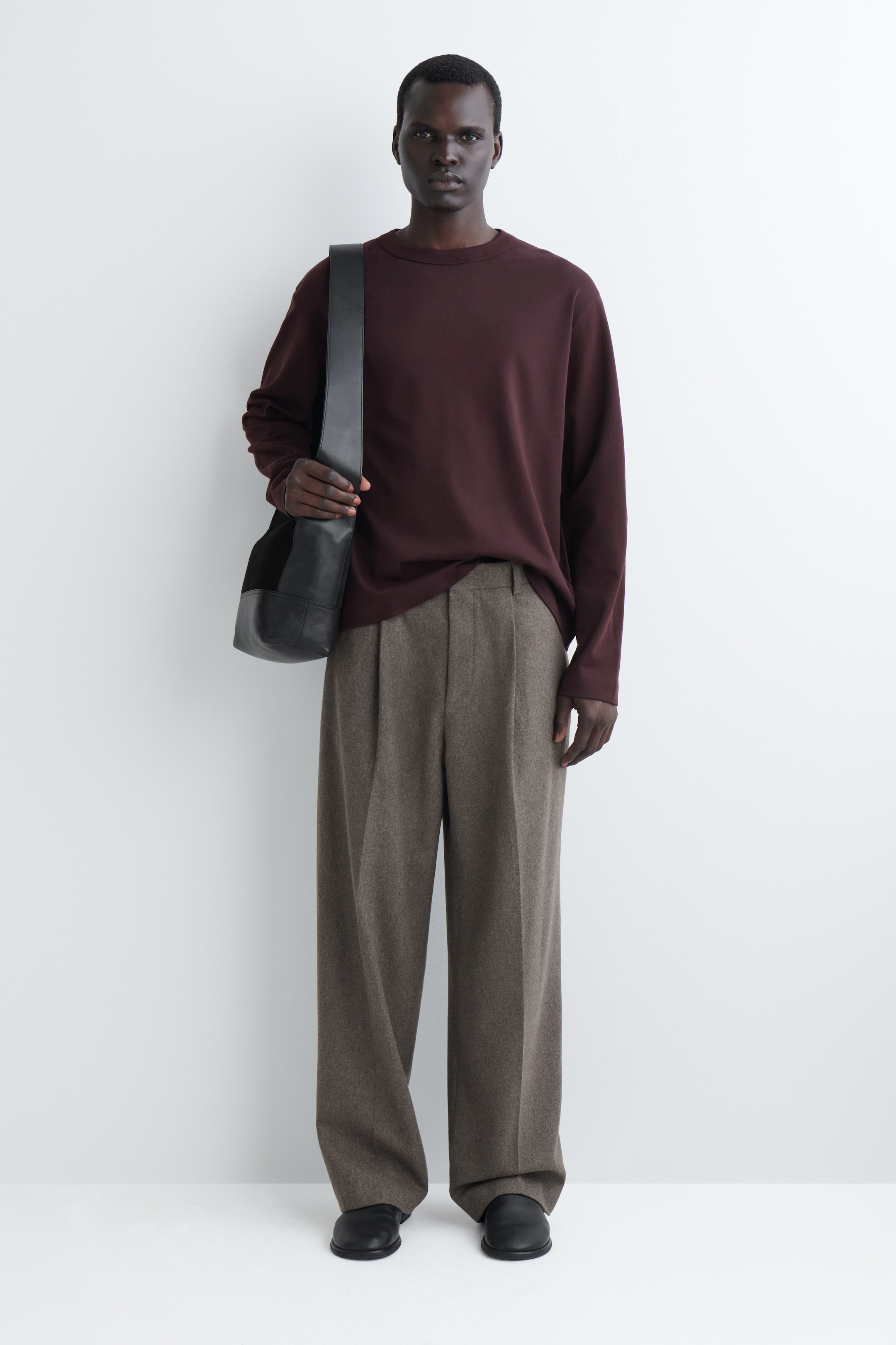 Ver imagen más grande: Un top burdeos de ajuste relajado, con cuello redondo y mangas largas está siendo usado, combinado con unos pantalones marrón jaspeado de pierna ancha y pliegues. Un bolso de hombro de cuero negro se lleva sobre el hombro izquierdo, descansando contra el top, y unos zapatos negros sin cordones son visibles en la parte inferior.