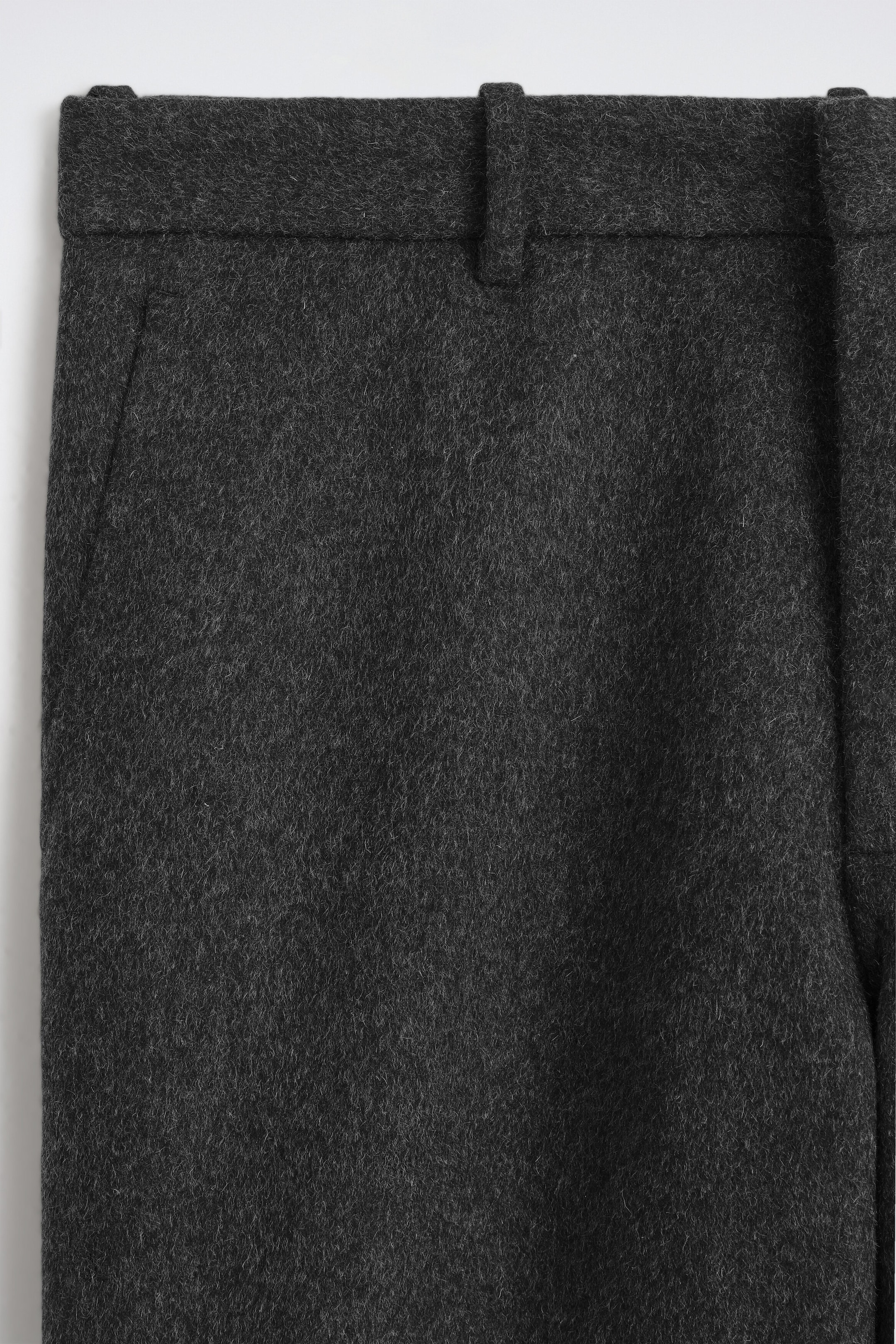 Dark grey wool-blend trousers are shown in a close-up front view, showcasing the soft, textured fabric, a structured waistband with belt loops, and a side slip pocket. Dark grey wool-blend trousers are shown in a close-up front view, showcasing the soft, textured fabric, a structured waistband with belt loops, and a side slip pocket.