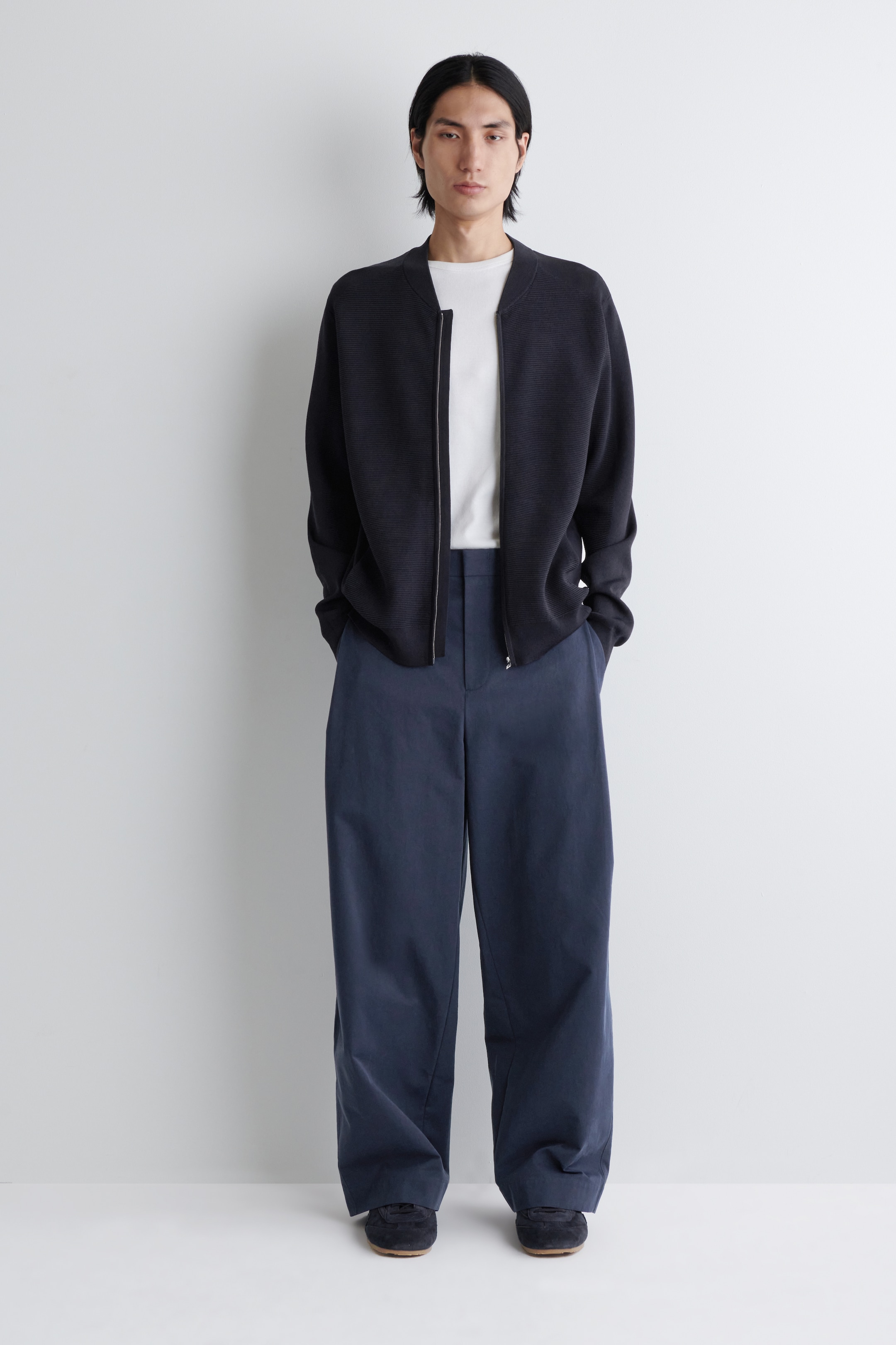 Visualizza immagine più grande: Una giacca bomber blu scuro con texture, caratterizzata da un colletto a fascia e una cerniera frontale color argento, è indossata sopra una t-shirt bianca con scollo a girocollo. L'outfit è completato da pantaloni a gamba larga blu scuro e scarpe in camoscio scuro, il tutto presentato da una vista frontale.