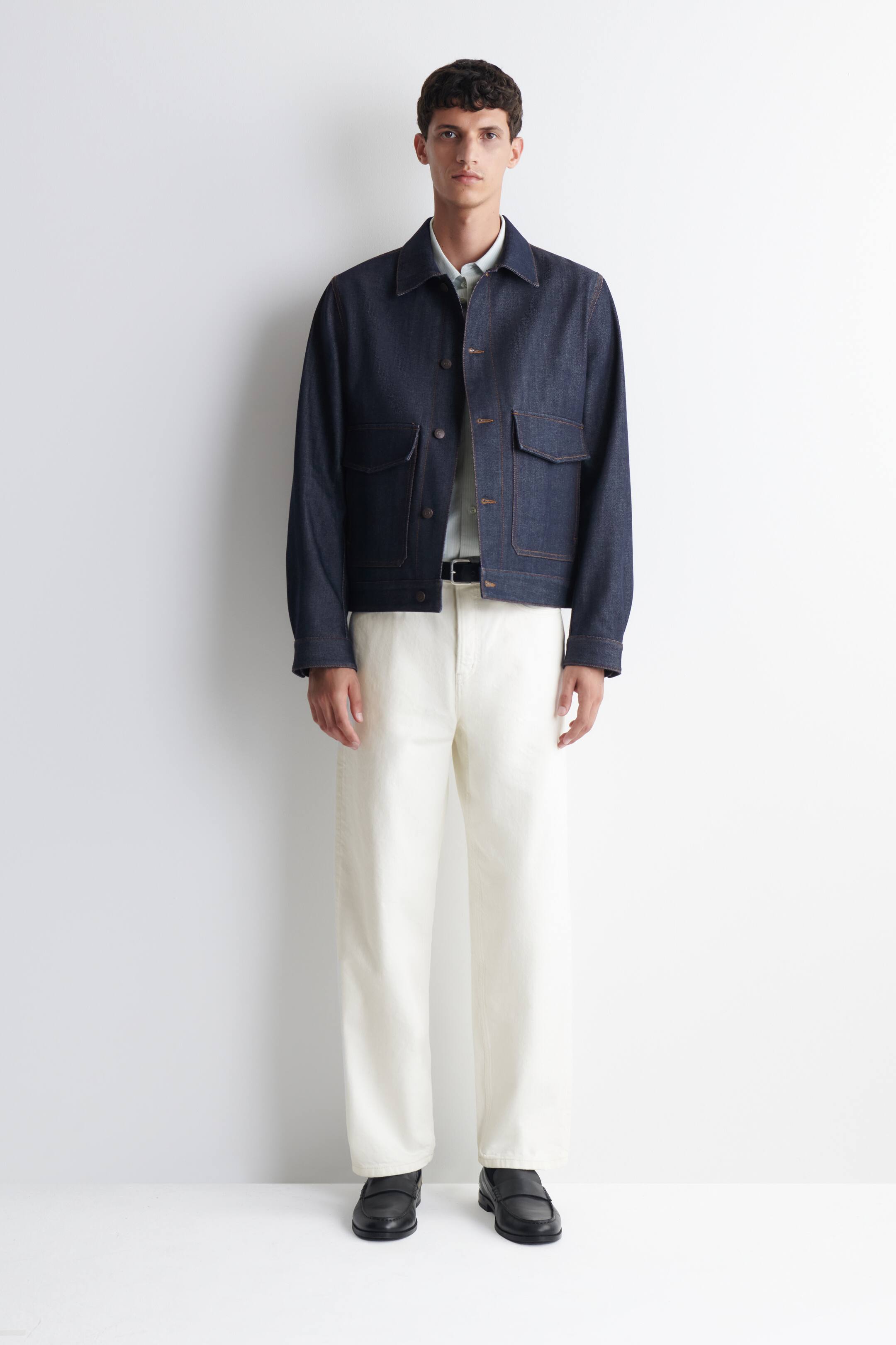 View larger image: A dark-wash denim jacket, featuring contrast stitching and four patch pockets, is layered over a light collared shirt. It is styled with light cream wide-leg trousers, a dark belt, and black leather loafers.