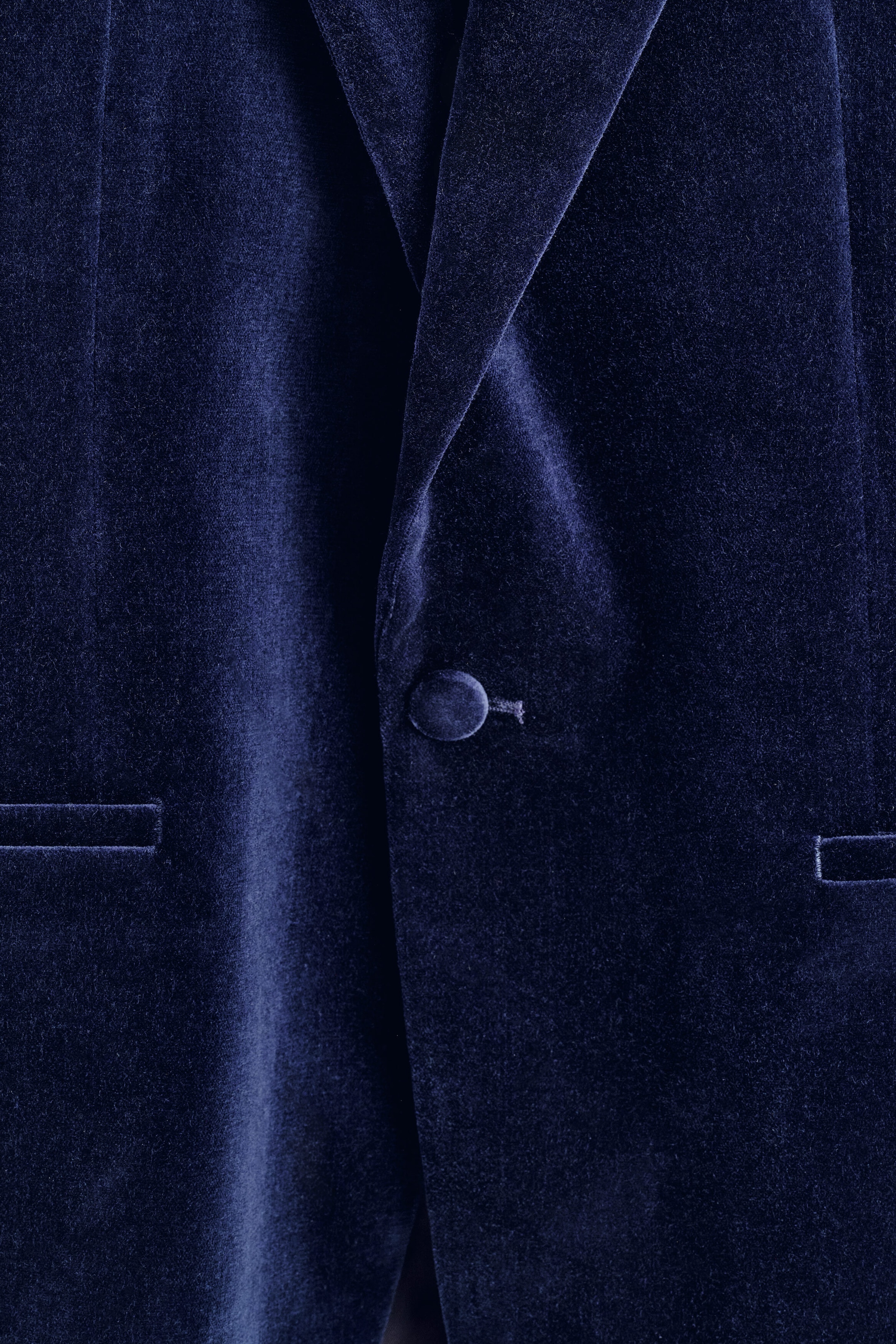A close-up front view of a navy blue velvet tuxedo blazer, highlighting the rich, plush texture of the fabric, a single-breasted closure with a fabric-covered button, and partially visible welt pockets. A close-up front view of a navy blue velvet tuxedo blazer, highlighting the rich, plush texture of the fabric, a single-breasted closure with a fabric-covered button, and partially visible welt pockets.