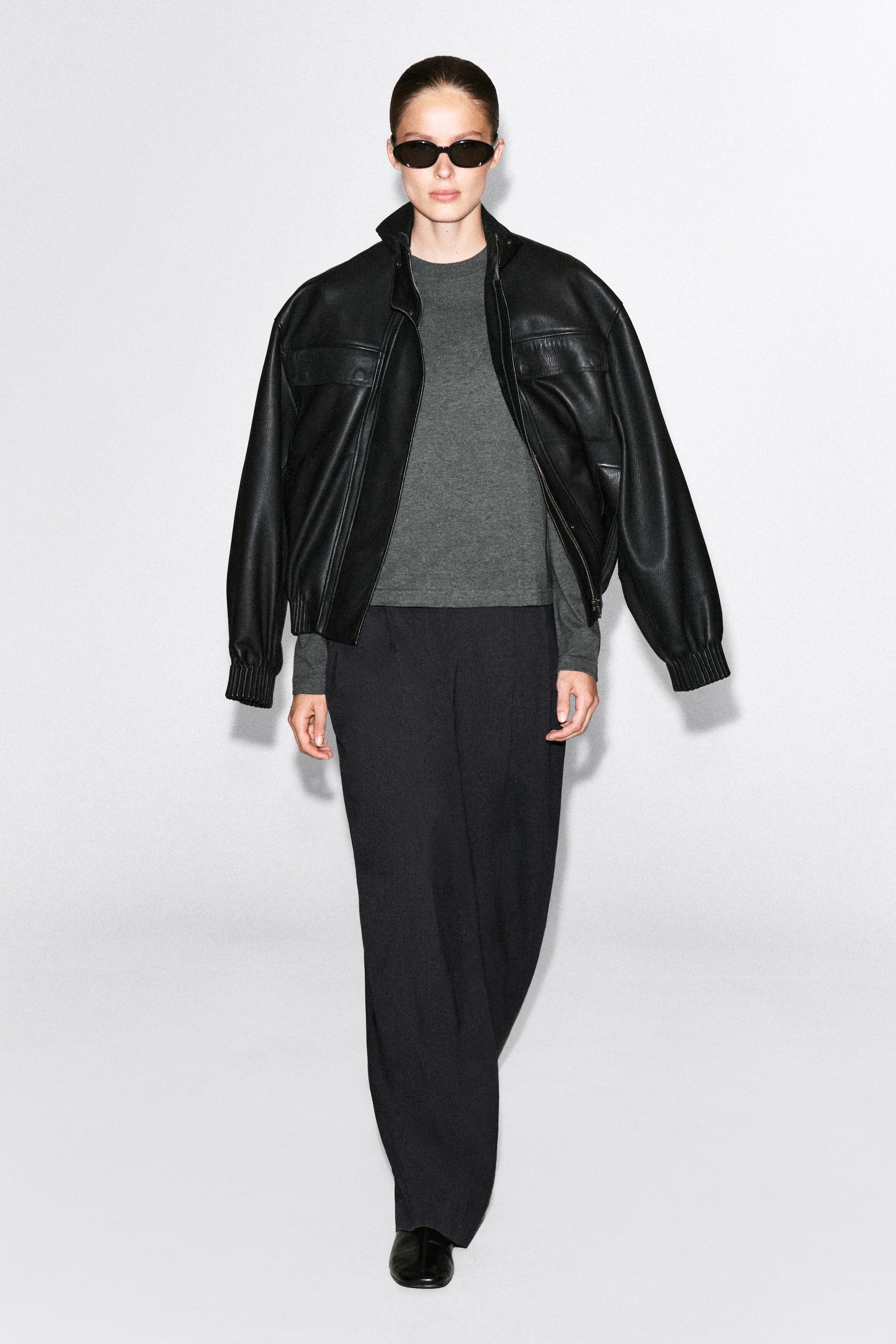 The model is wearing a gray, long-sleeved crewneck T-shirt layered under an open black leather jacket. The T-shirt features a clean-cut hem and is paired with dark, wide-leg trousers.