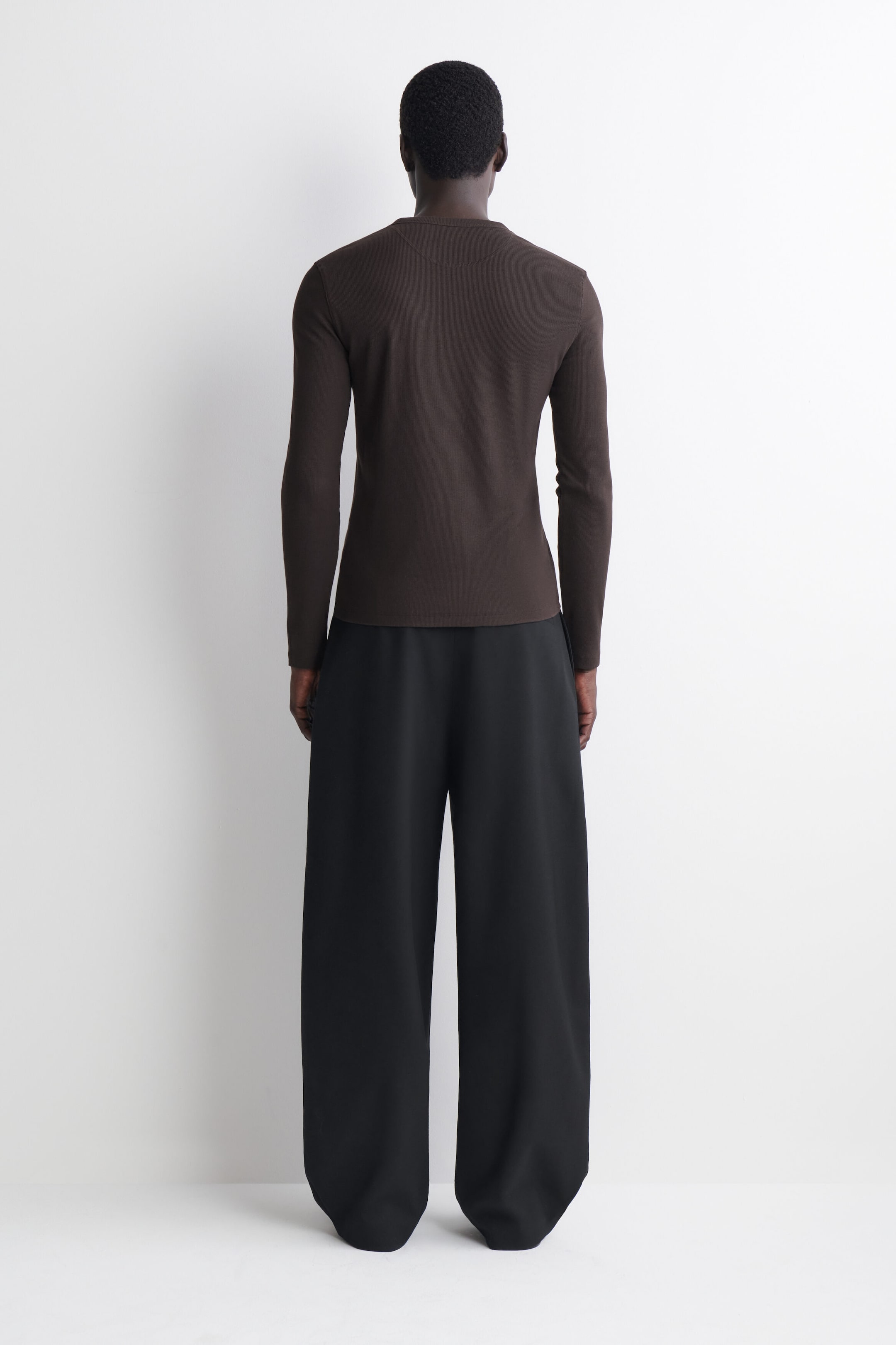 Ciemnobrązowy, dopasowany longsleeve Henley z prążkowanej bawełny jest prezentowany na modelu tyłem, co podkreśla jego przylegający do sylwetki krój, prosty dół oraz długie rękawy. Koszulkę zestawiono ze spodniami o szerokich nogawkach w kolorze czarnym, pokazując, jak dół układa się na górnej części bioder. Ciemnobrązowy, dopasowany longsleeve Henley z prążkowanej bawełny jest prezentowany na modelu tyłem, co podkreśla jego przylegający do sylwetki krój, prosty dół oraz długie rękawy. Koszulkę zestawiono ze spodniami o szerokich nogawkach w kolorze czarnym, pokazując, jak dół układa się na górnej części bioder.