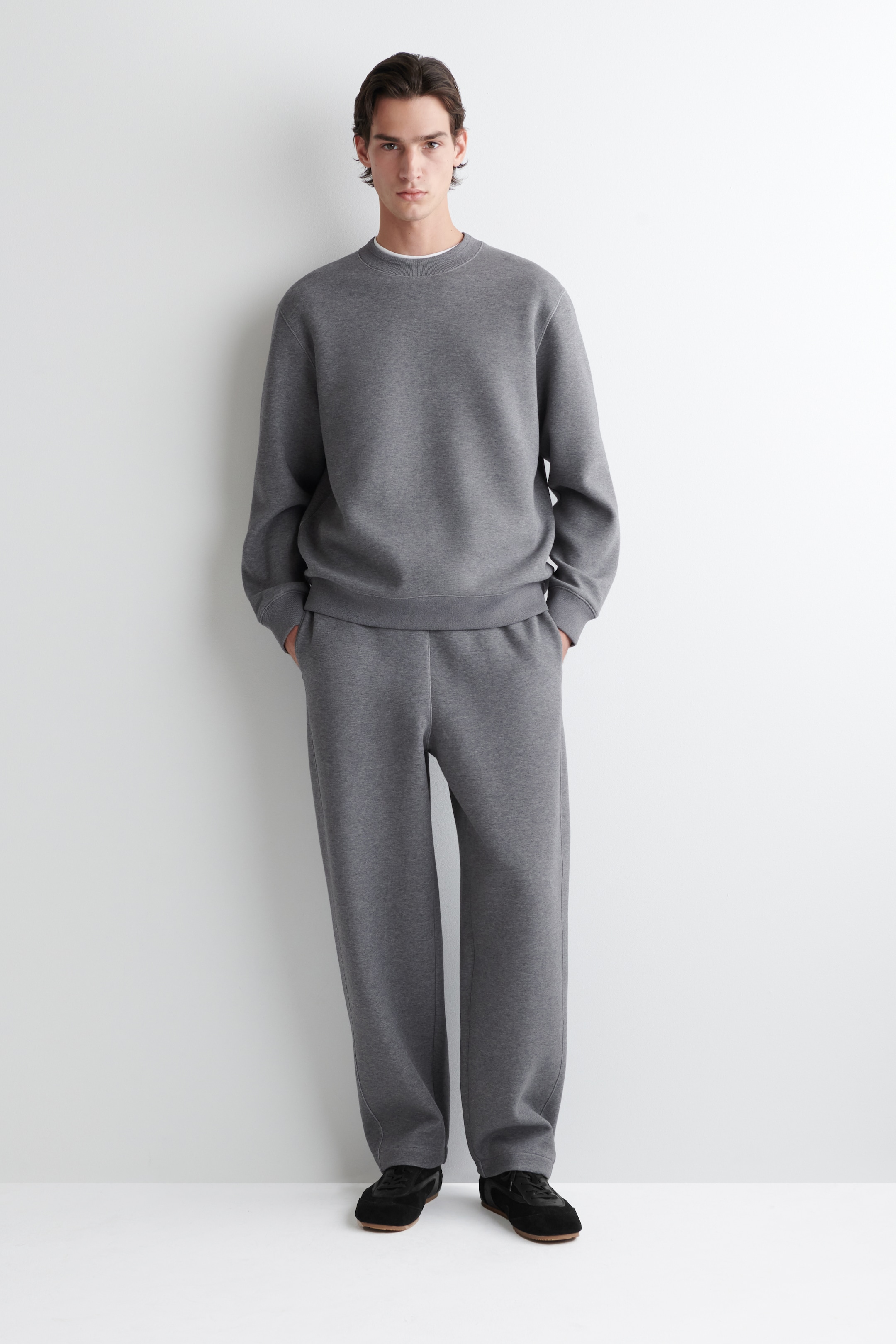 Visualizza immagine più grande: Una felpa girocollo grigio scuro e pantaloni della tuta a vestibilità rilassata abbinati sono presentati su un modello rivolto in avanti. La felpa rivela un colletto di t-shirt bianca sotto, e i pantaloni della tuta presentano tasche laterali.
