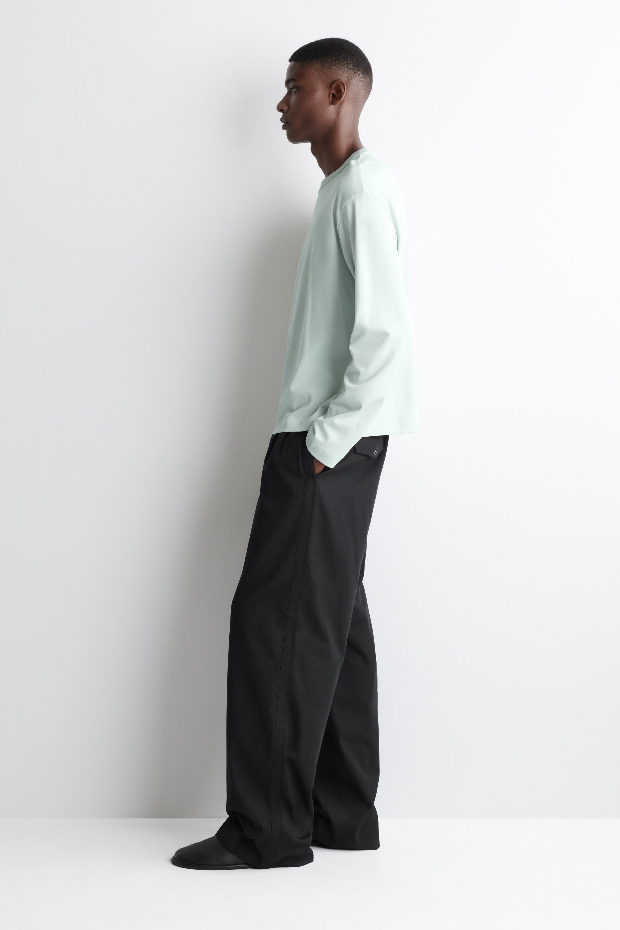 View larger image: A loose-fitting light green long-sleeved top is being worn with black wide-leg trousers, and the ensemble is being presented from a side angle. The top is showing a simple crew neck, while the trousers are displaying a side pocket and a small buttoned flap pocket on the thigh, and are draping over black shoes.