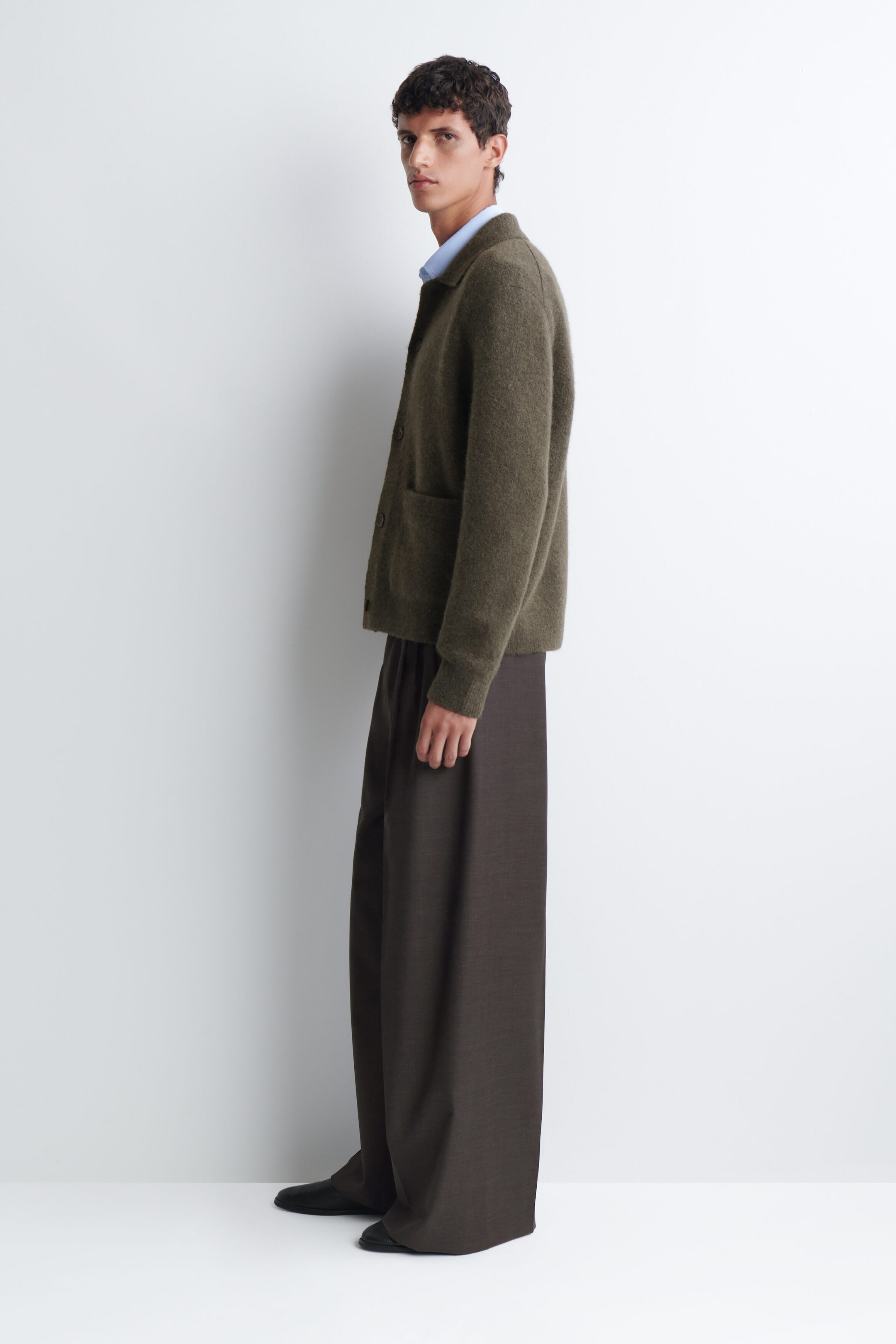 A textured collared cardigan in olive green is being worn by the model, featuring a fuzzy texture, a shirt-style collar, button closures, and a side patch pocket. The cardigan is layered over a light blue collared shirt and styled with dark, wide-leg trousers. A textured collared cardigan in olive green is being worn by the model, featuring a fuzzy texture, a shirt-style collar, button closures, and a side patch pocket. The cardigan is layered over a light blue collared shirt and styled with dark, wide-leg trousers.