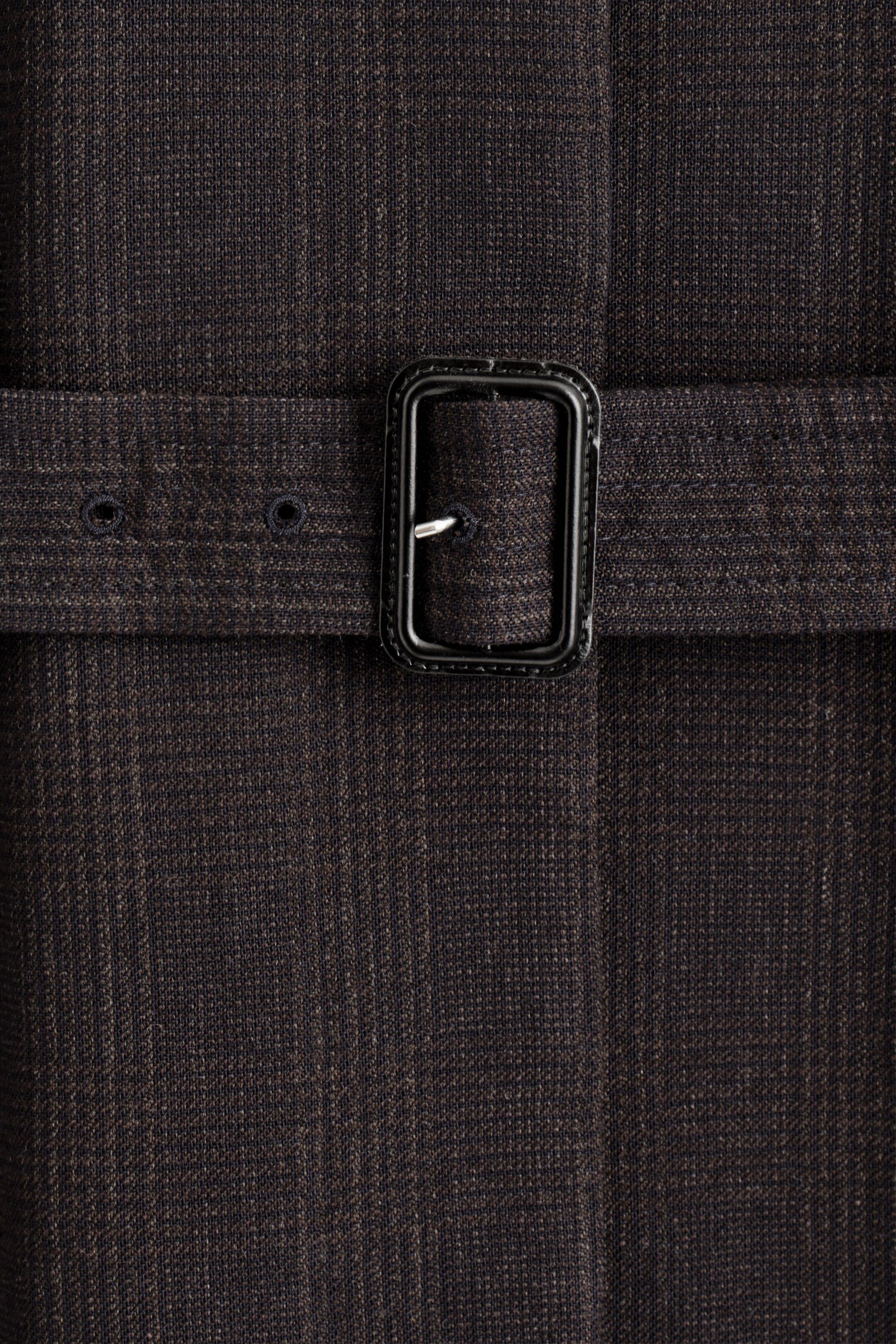 Agrandir l'image: Un gros plan d'un trench-coat marron foncé montre une texture subtile de rayures verticales sur son tissu. Une ceinture en tissu assortie s'étend sur le devant du manteau, fixée par une boucle rectangulaire en métal foncé avec une seule ardillon et deux œillets visibles.